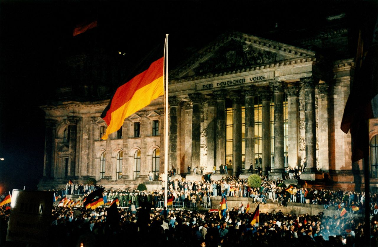 Menschenmenge vor dem Reichstag bei Nacht. Vorne und zentral groß im Bild: eine gehisste Deutschlandfahne.