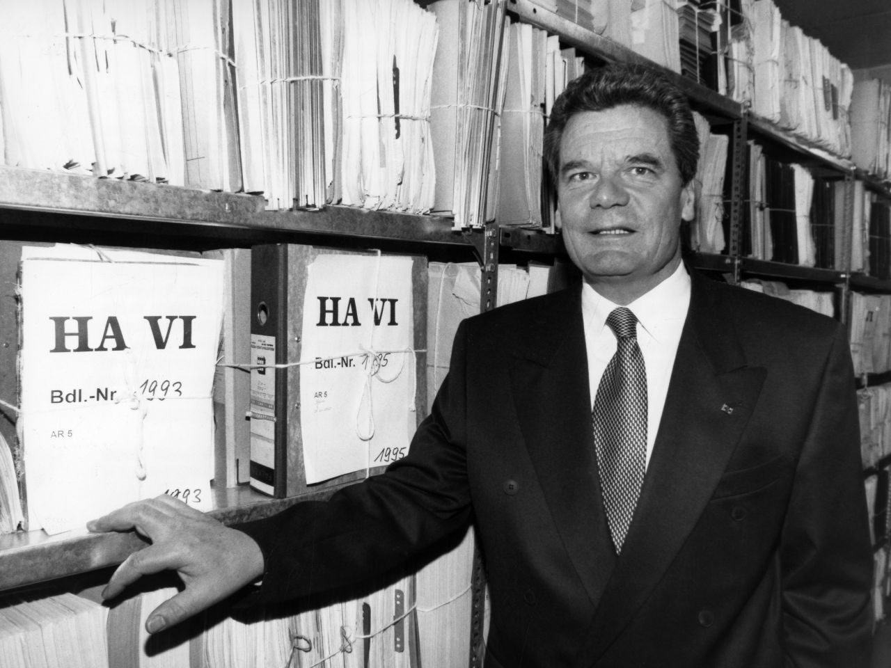 schwarz-weiß Fotografie, Joachim Guack steht vor einem Archivregal, das übervoll it Akten ist. Auf zwei Aktendeckeln ist zu lesen: HA VI. Gauck fast mit der einen Hand ans Regal und schaut freundlich aber unsicher mit geöffnetem Mund in die Kamera.