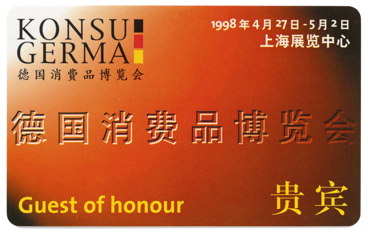 Rechteckige Karte; Vorderseite: orangegrundig, zu den Rändern dunkler werdend, links oben schwarz-rot-goldenes Logo sowie: Konsu / Germa; über Karte verteilte chinesische Schriftzeichen, links unten in Gelb: Guest of Honour; Rückseite: graugrundig, weißes Unterschriftsfeld.