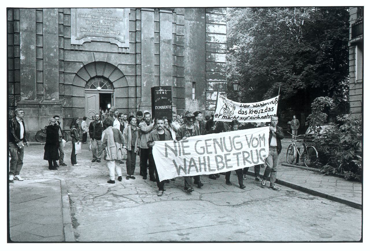 Abzug wahrscheinlich 1998, leichte Schrägansicht vom linken Bürgersteig auf den Demonstrationszug, der sich vor der Sophienkirche formiert, Die Demonstranten tragen Transparente wie NIE GENUG VOM / WAHLBETRUG und Der Wähler muß / das Kreuz, das / er macht, auf sich nehmen, 2 Männer in der 2. Reihe tragen einen schwarzen Kasten mit der weißen Aufschrift URNE / HIER RUHT DIE / DEMOKRATIE, im HG Eingang der Sophienkirche, aus der weitere Personen folgen, der Eingang trägt im historischen Supraport die Aufschrift Wie lieblich / sind Deine Wohnungen / Herr Zebaoth ! / Höre des Herrn Wort!