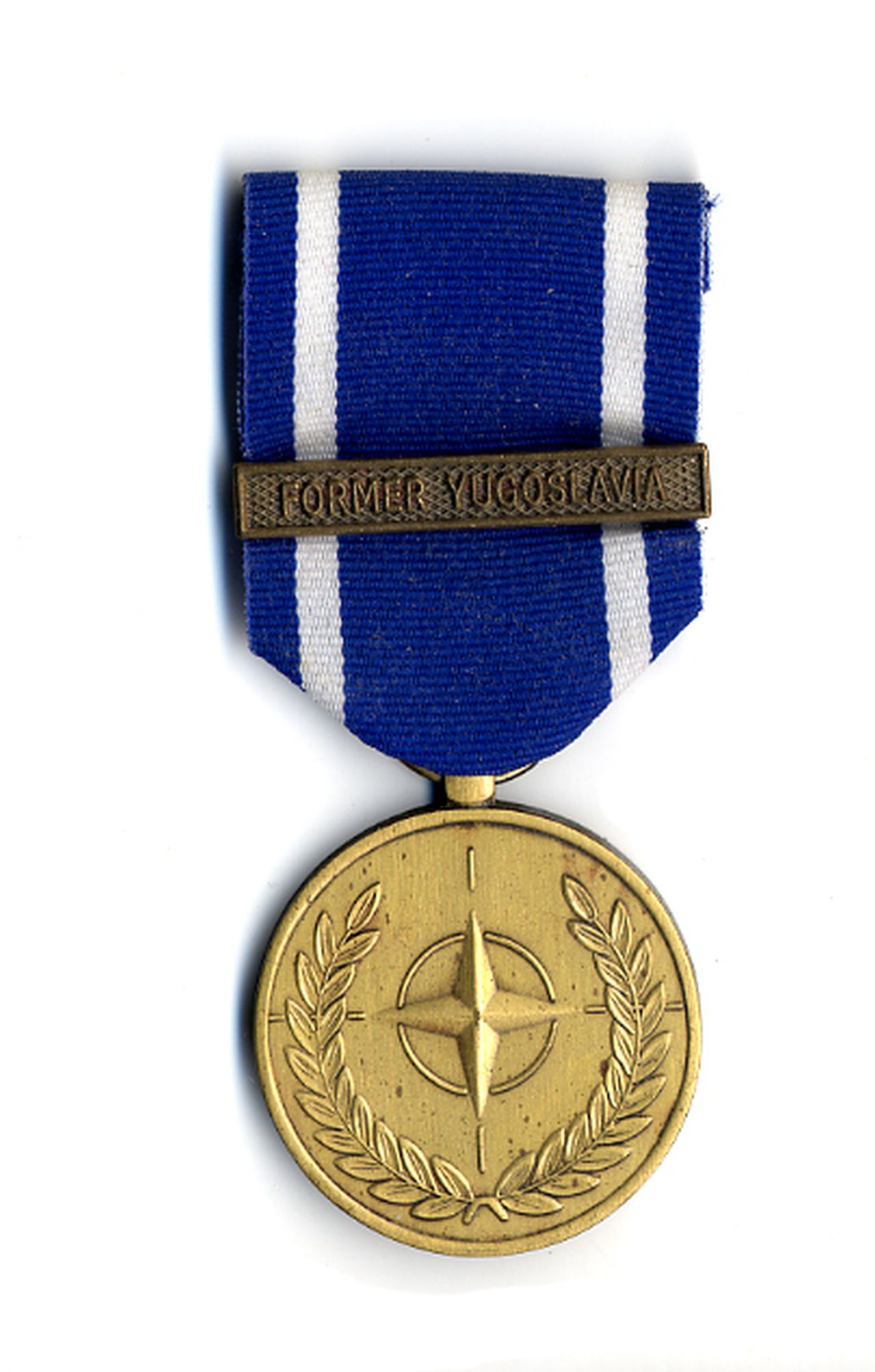 Goldfarbene Medaille mit Randstab. Erhabener Lorbeerkranz außen. Im Zentrum erhabenes NATO-Emblem. Rückseite: Randstab. Englisch-französische Umschrift: North Atlantic Treaty Organization. Im Zentrum zweisprachig: In Service of Peace / and Freedom. Oben Öse mit Bandring. Durchgezogenes blaues Band mit schmalen weißen Randstreifen. Auf dem Band bronzefarbene Spange mit dem erhabenen Schriftzug Former Yugoslavia vor gekörntem Hintergrund.