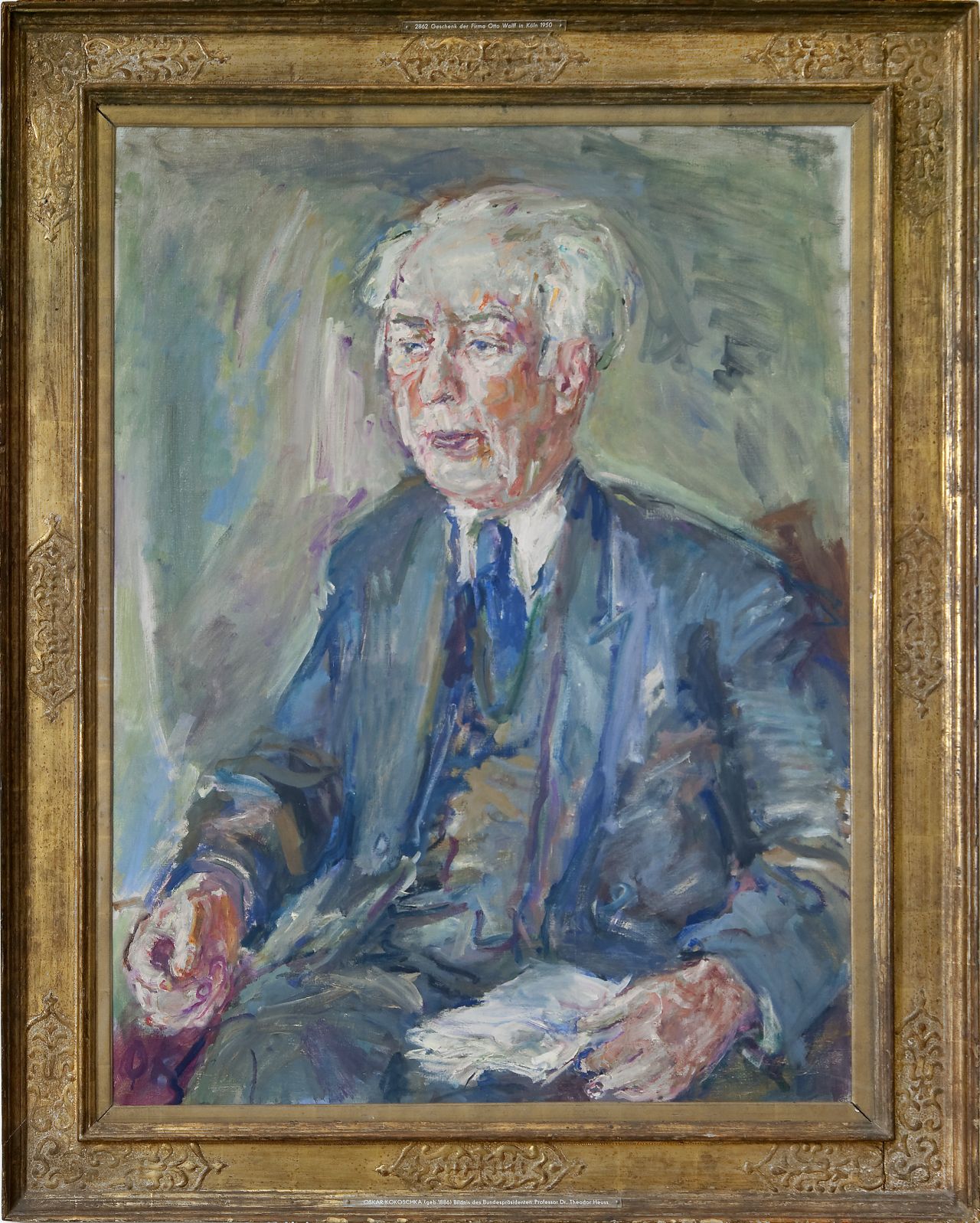 Das farbige Ölgemälde zeigt Theodor Heuss, sitzend, bis in Kniehöhe. Das Gemälde befindet sich in einem goldfarbenen Rahmen. Auf einem Schild unten steht: 'Oskar Kokoschka (geb. 1886) Bildnis des Bundespräsidenten Professor Dr. Theodor Heuss'.