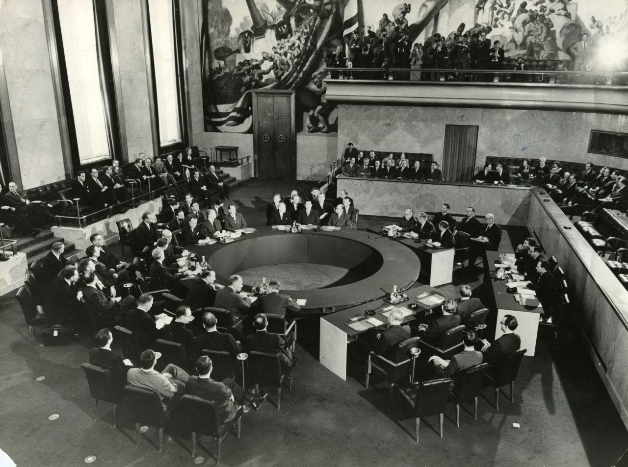 Teilnehmer der Konferenz sitzen in Delegationen um einen ringförmigen Verhandlungstisch. Im Hintergrund sitzen auf einer rechteckig darum geformten, erhöhten Galerie weitere Konferenzteilnehmer.