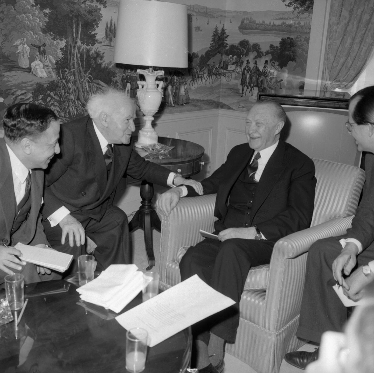 Rechts in einem Sessel sitzt lächelnd Adenauer in einem Sessel. Über Eck ihm zugewandt redend und Adenauers Unterarm berührend sitzt Ben-Gurion. Eine weitere Person sitzt links neben Ben-Gurion. Vor ihnen ein Tisch mit Gläsern und Papieren darauf.