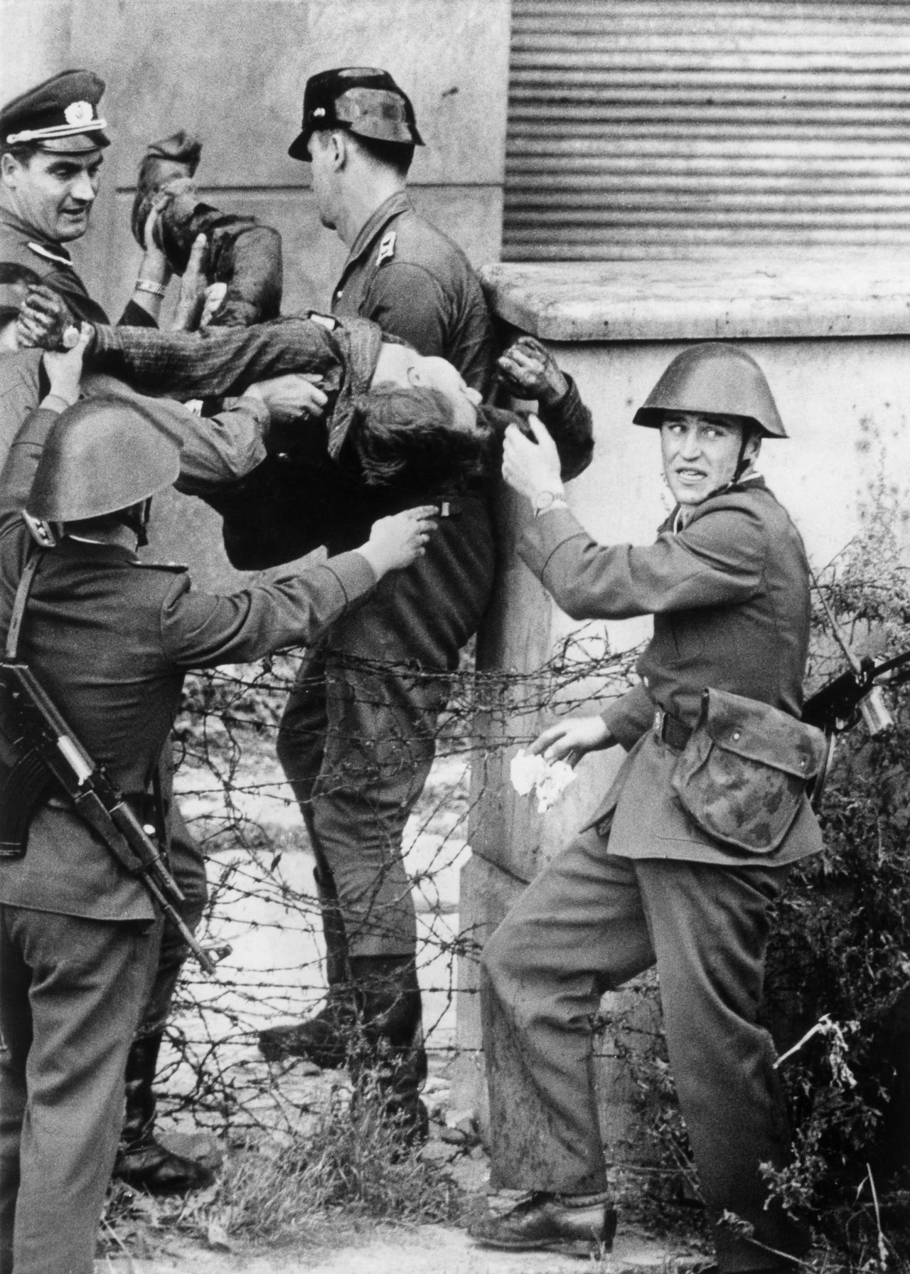 Schwarzweiß-Aufnahme: vier DDR-Grenzsoldaten bergen den leblosen Peter Fechter nach dessen gescheiterter Flucht; zwischen den Grenzern Stacheldraht, im Hintergrund eine Hausfassade.