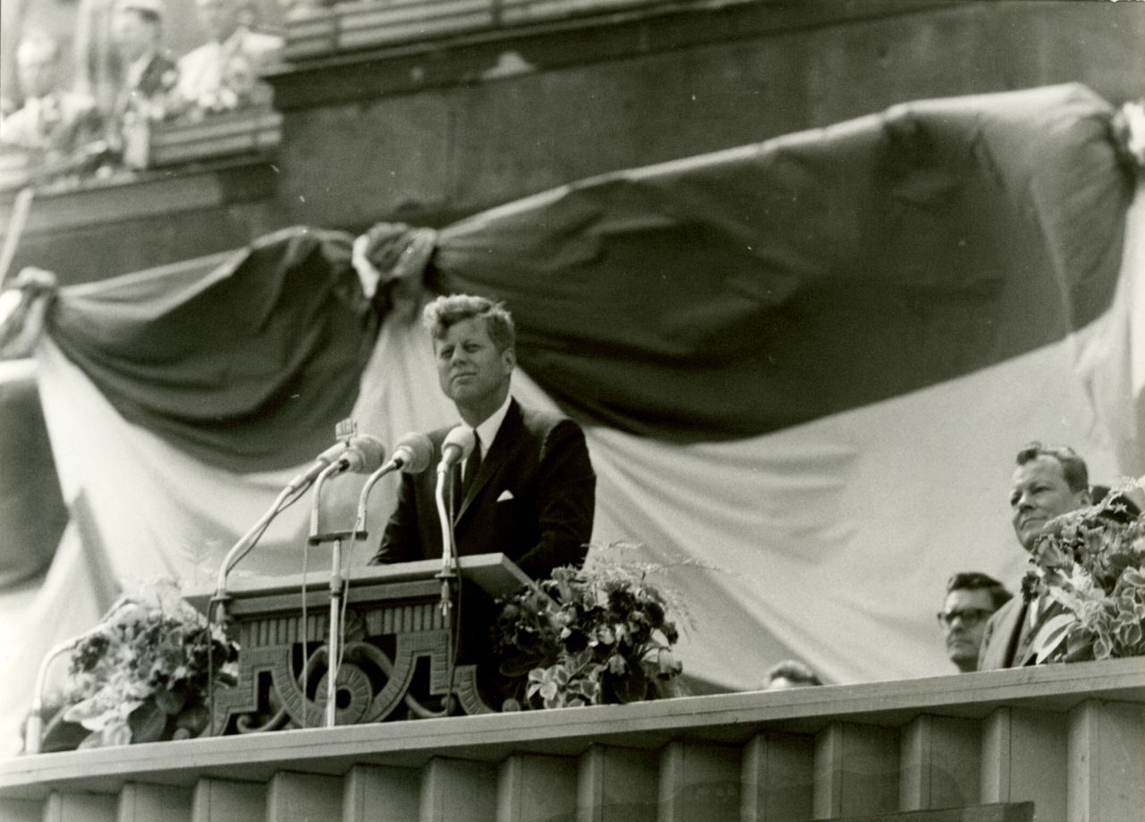 US-Präsident Kennedy hinter einem Rednerpult vor dem Schöneberger Rathaus. Auf dem von Blumen umrahmten Pult mehrere Mikrofone. Etwas entfernt rechts steht Willy Brandt. Hinter ihnen der mit Fahnen abgehangene Vorbau des Rathauses.