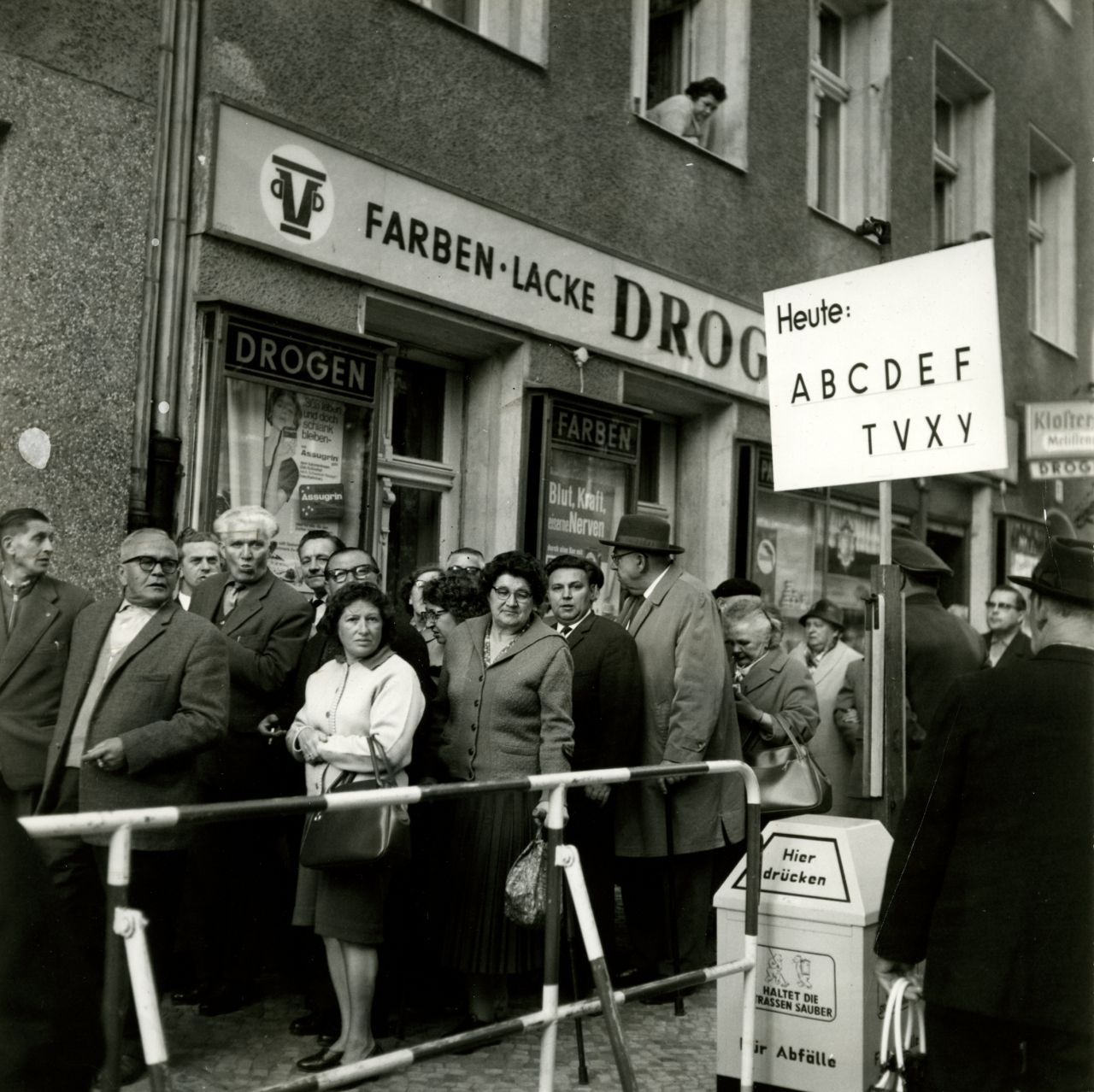 Warteschlange vor Passierstelle;  im Vordergrund eine Absperrung , ein Abfalleimer und ein Schild mit der Aufschrift ‚Heute: A B C D E F T V X Y‘. Im Hintergrund eine Häuserfassade mit Drogerie; darüber eine Frau, die aus dem Fenster nach unten schaut.