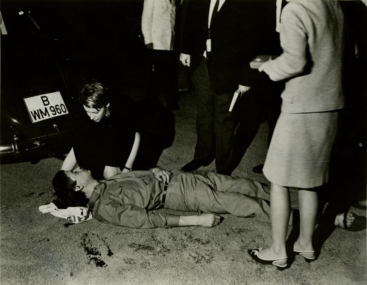 Benno Ohnesorg liegt schwer verletzt auf dem Rücken am Boden; unter seinem Kopf ein Tuch. Eine Frau an seinem Kopf kniend beugt sich über ihn. Zwei weitere Personen stehen um ihn herum. Im Hintergrund das Heck eines Autos mit dem Kennzeichen ‚B WM 960‘. 