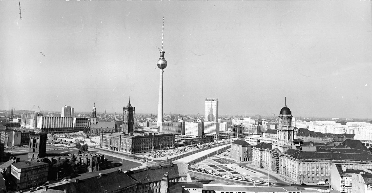 Schwarzweiß-Stadtansicht von Ost-Berlin von erhöhter Position aus. Im Zentrum Ost-Berliner Fernsehturm. Davor Straßenkreuzung. Im Vordergrund ältere Gebäude teils mit Türmen, direkt hinter und neben dem Fernsehturm neuere mehrstöckige Gebäude und Hochhäuser. 