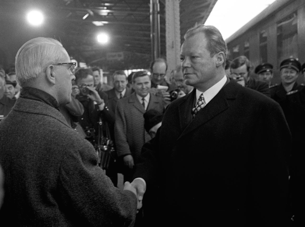 Willy Brandt, rechts im Bild, schüttelt Willi Stoph, links im Bild, die Hand. Brandts Gesichtsausdruck ist neutral, Stophs Gesicht ist nicht zu sehen. Im Hintergrund stehen Personen, die die Szene fotografieren oder beobachten.