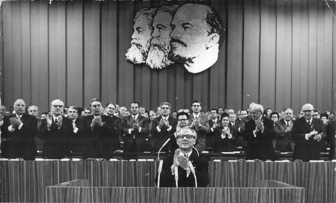 Im Vordergrund Erich Honecker applaudierend am Rednerpult mit Mikrofonen. Dahinter höher stehend Präsidium und Delegierte, ebenso Beifall klatschend. An der Wand hinter ihnen ein Porträtbild von Karl Marx, Friedrich Engels und Wladimir Iljitsch Lenin.