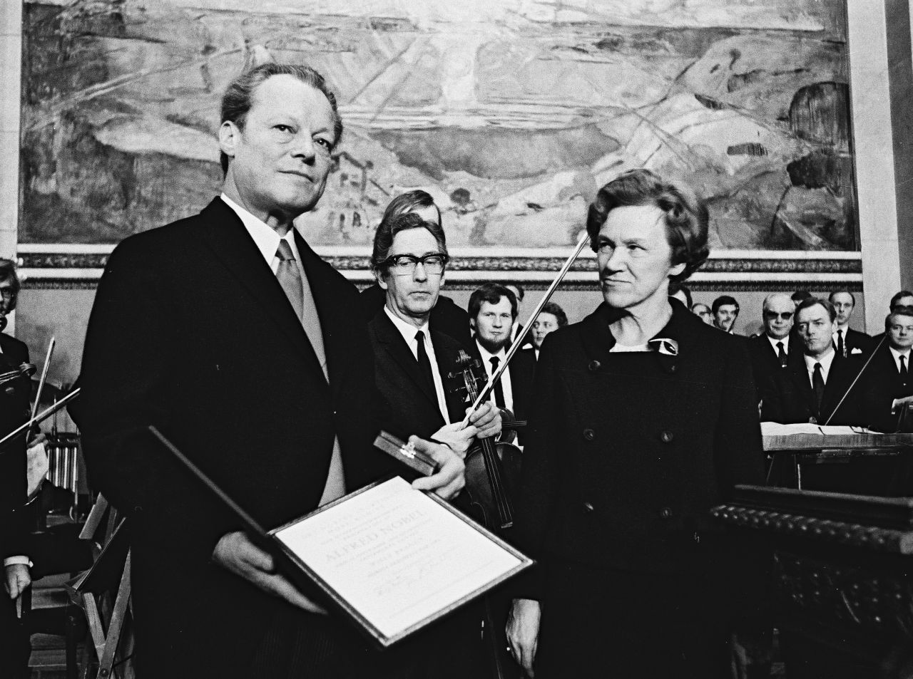 Willy Brandt, links im Bild, hält den Friedensnobelpreis, d.h. eine Urkunde, und ein Kästchen in den Händen und schaut über die Kamera hinweg. Neben ihm steht Aase Lionæs, die ihn anschaut. Im Hintergrund steht ein Orchester.
