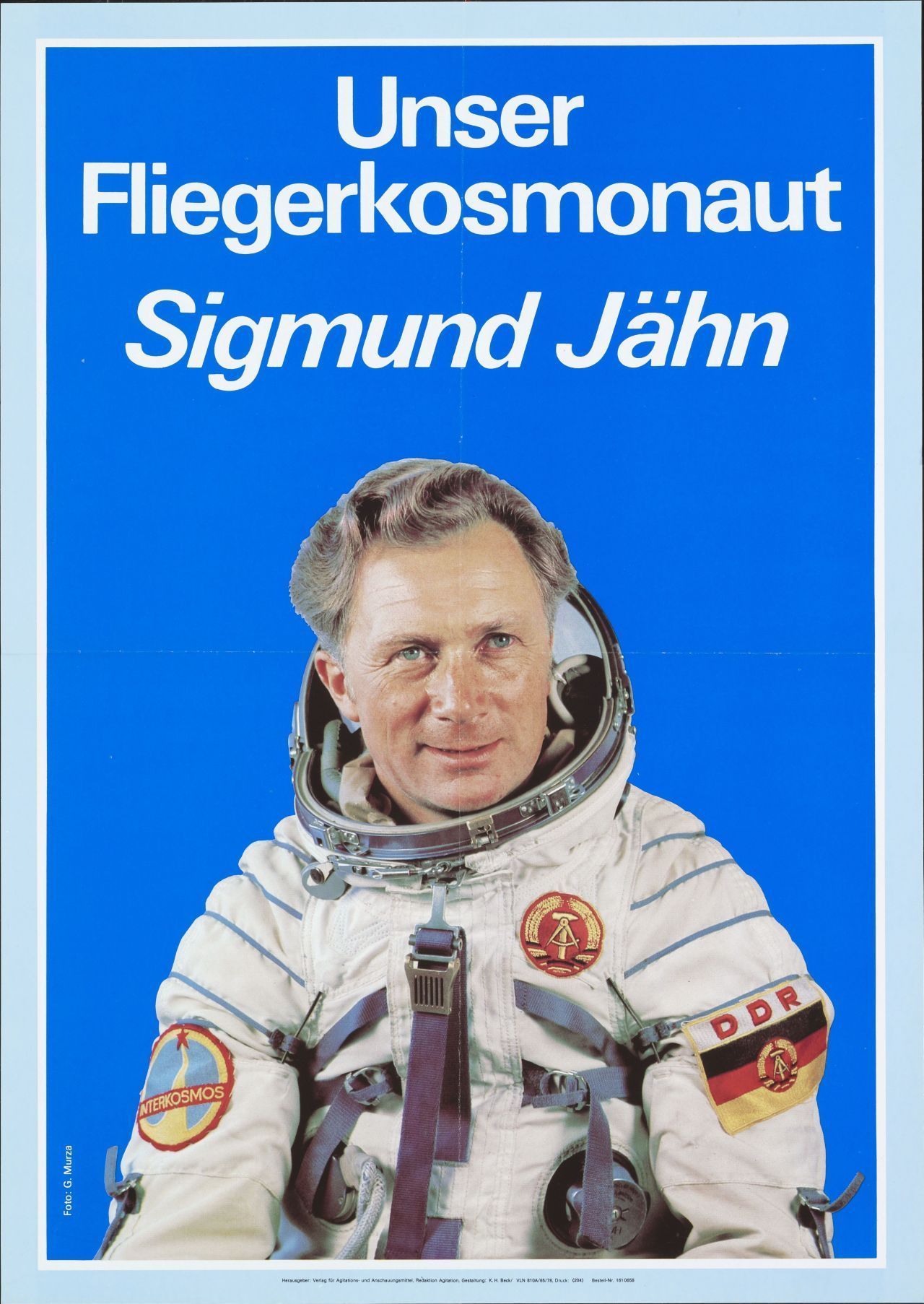 Hochformatiges Plakat, vor leuchtend blauem Hintergrund das freigestellte Halbfigurenporträt als Foto von Sigmund Jähn. Er trägt einen Raumanzug. Oben weißer Schriftzug: ‚Unser Fliegerkosmonaut Sigmund Jähn‘. Drum herum schmaler hellblau-weißer Rahmen.