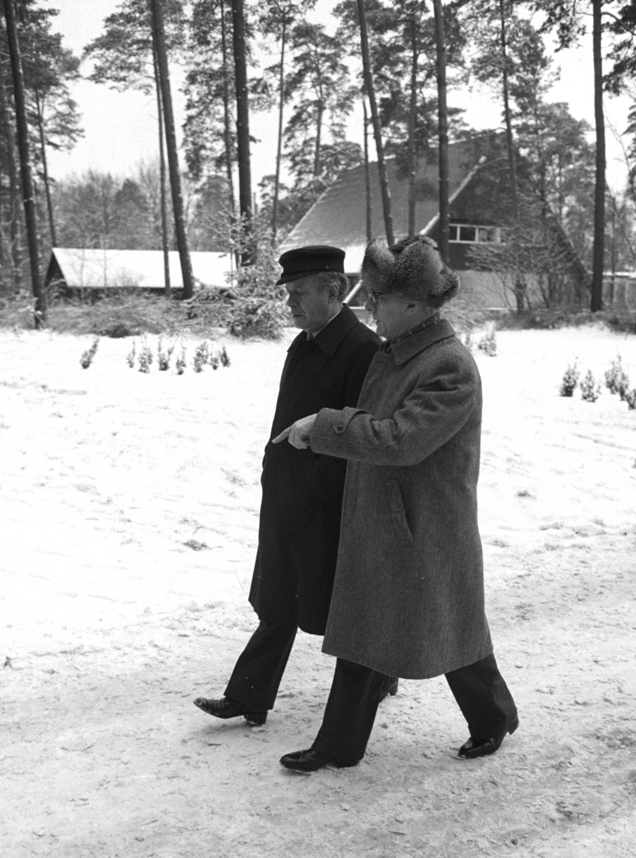 Schwarzweiß-Foto von Helmut Schmidt und Erich Honecker im Gespräch bei einem Spaziergang in schneebedeckter Landschaft. Beide tragen Mäntel, Honecker eine Pelzmütze und Schmidt eine Elblotsenmütze. Im Hintergrund ein von Bäumen umgebenes Haus.