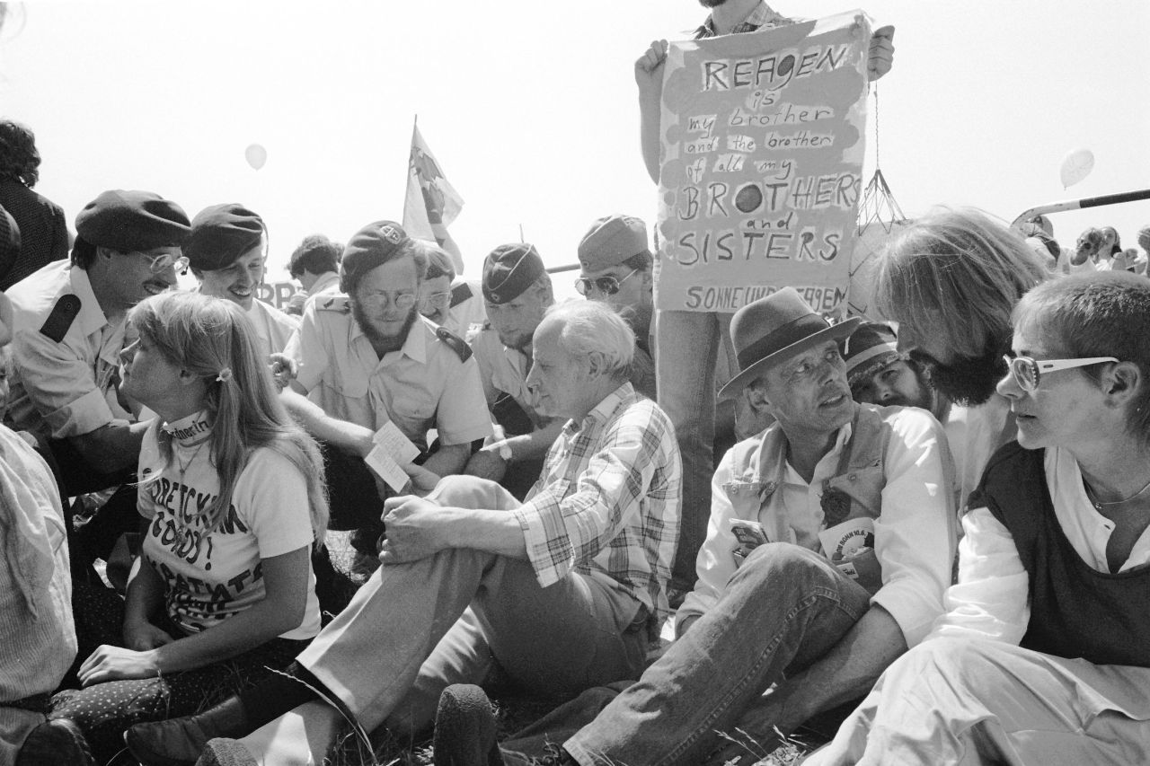 Schwarzweiß-Foto, sitztende Demonstranten. Links Gert Bastian, im Gespräch mit Polizisten. Rechts davon Joseph Beuys, spricht mit Lukas Beckmann. Hinter ihnen Person mit Plakat, darauf: ‚Reagan is my brother and the brother of all my BROTHERS and SISTERS‘.