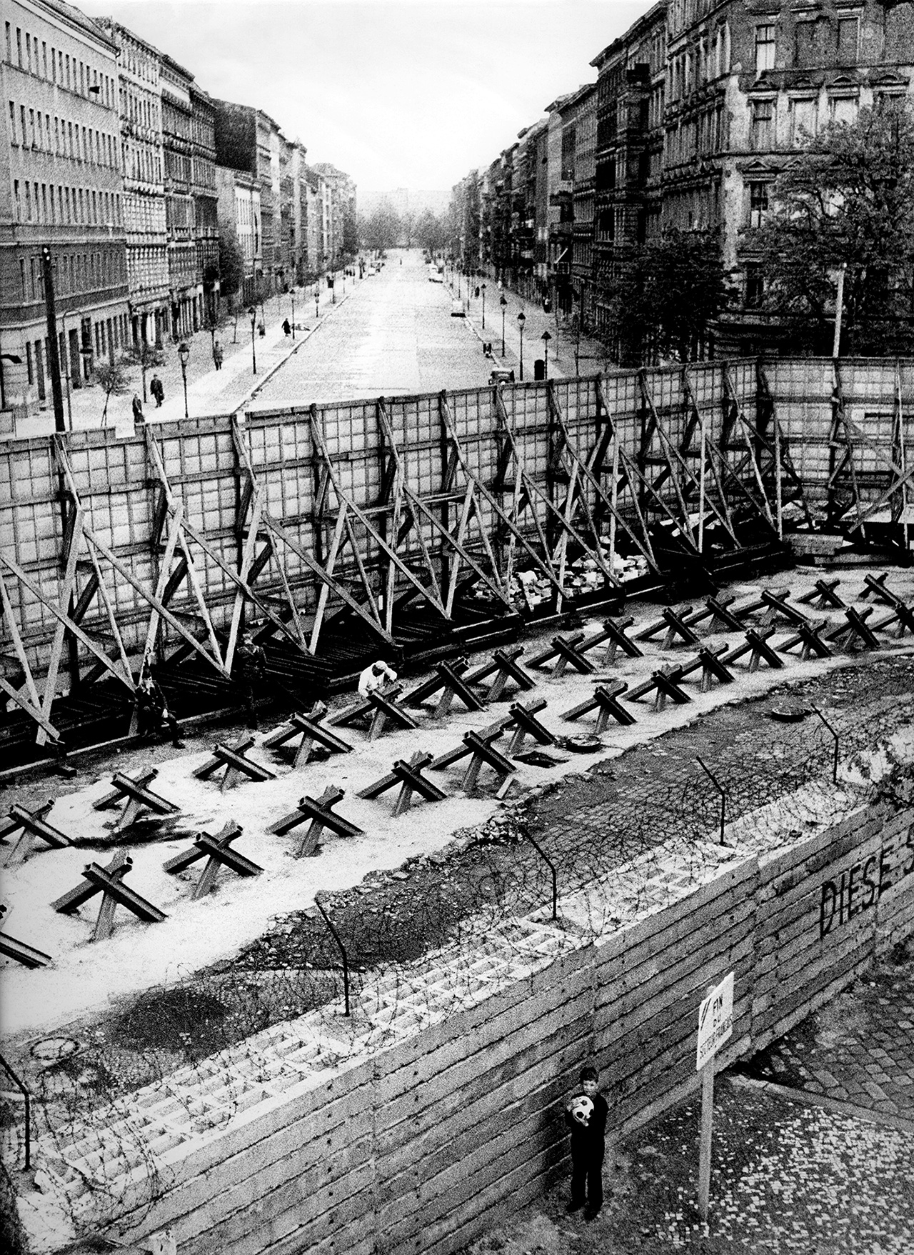 Der Bau der Berliner Mauer zementiert die Teilung Deutschlands und Berlins endgültig. 
An vielen Stellen der Stadt werden Straßen plötzlich zu Sackgassen, Häuser stehen direkt auf der Grenze. Besonders deutlich wird dies an der Bernauer Straße, die die Grenze zwischen den Stadtbezirken Mitte – im Osten – und Wedding – im Westen – markiert. Die Häuser auf der Ost-Seite der Bernauer Straße werden im Zuge des Mauerbaus abgerissen, und müssen Grenzanlagenweichen.
Der Fotograf Günter Zint fotografiert 1963 aus West-Berliner Perspektive die Grenze an der Bernauer Straße: Olaf, der kleine West-Berliner Junge auf dem Bild, wohnt im Wedding und kommt oft an die Mauer, um hier zu spielen. Auf der anderen Seite der Mauer ist gleichzeitig der Todessteifen zu sehen, der von Grenzsoldaten bewacht und von Bauerarbeitern ausgebaut wird.
