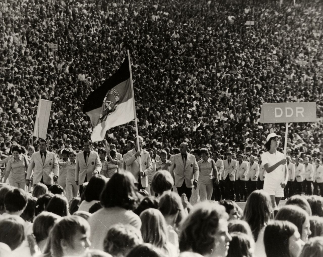 Mittelgrund: Eine Formation von Männern in Anzügen und Frauen in Blusen. An ihrem Anfang trägt ein Mann die Flagge der DDR. Vor ihm geht eine junge Frau mit einem Schild mit der Aufschrift DDR. Im Hintergrund eine dicht besetzte Zuschauertribüne.