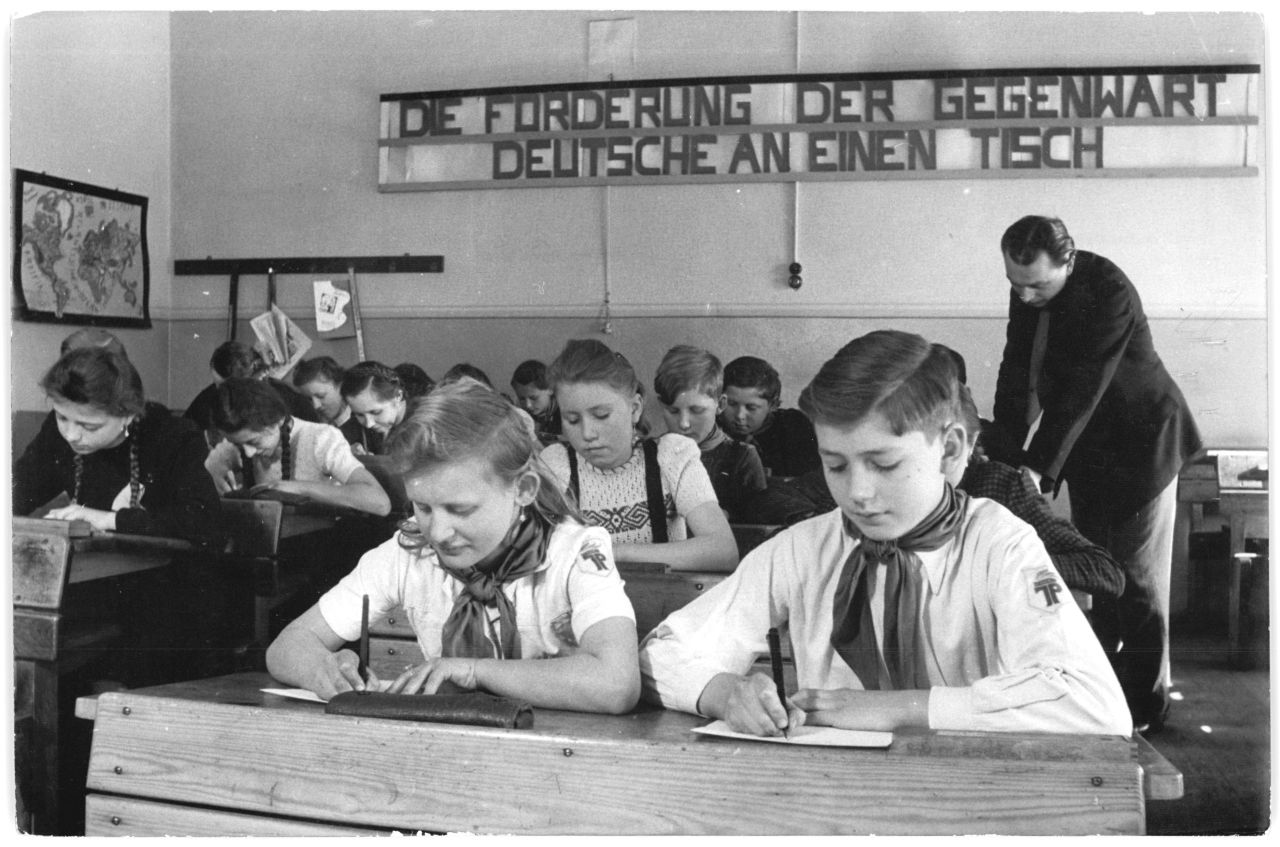 Schwarz/weiß-Fotografie: Junge Pioniere in der Schule beim Briefe schreiben. Im Hintergrund der Schriftzug 'Die Forderung der Gegenwart - Deutsche an einen Tisch'.