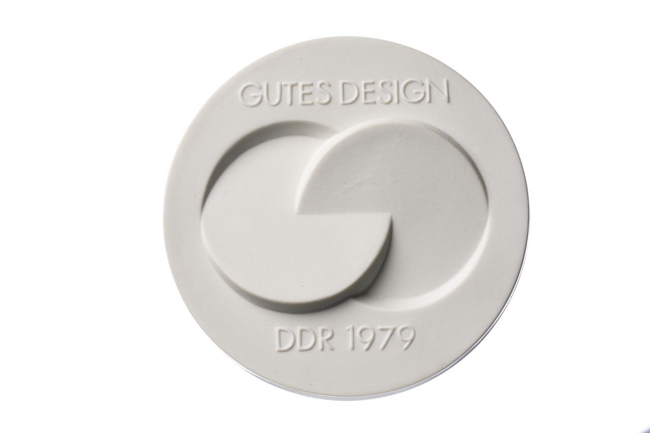 Weiße Porzellanmedaille, Vorderseite mittig erhabenes Signet, oben Gutes Design, unten DDR 1979, Rückseite DDR-Staatssymbol sowie Schwertermarke der Meißner Porzellanmanufaktur, weißes quadratisches Etui, innen mit weißem und blauem Stoff bezogen.