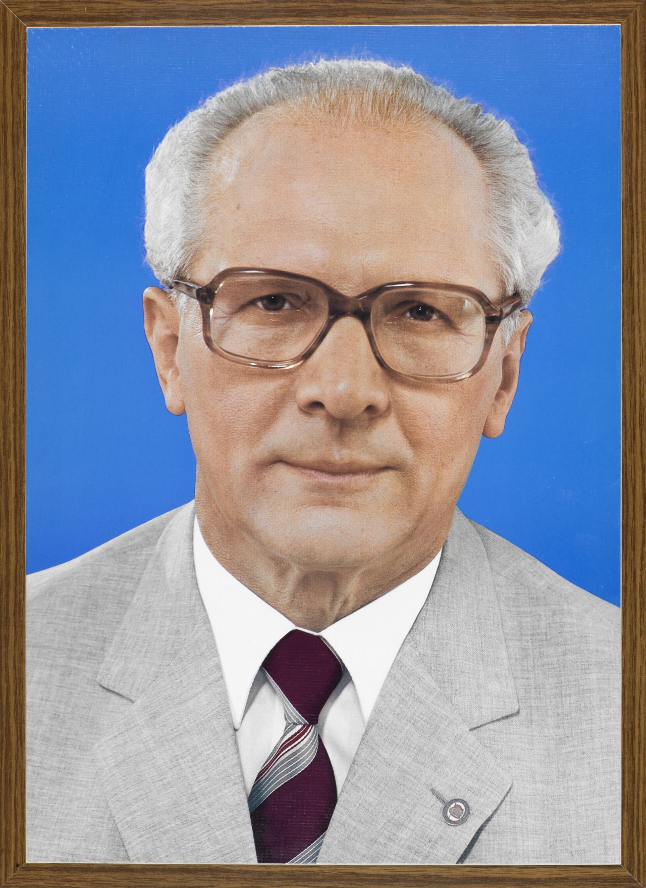 Ein Viertelporträt von Erich Honecker im hellgrauen Jacket mit SED-Anstecknadel, weißem Hemd und rot-grauer Krawatte. Er trägt eine Hornbrille.