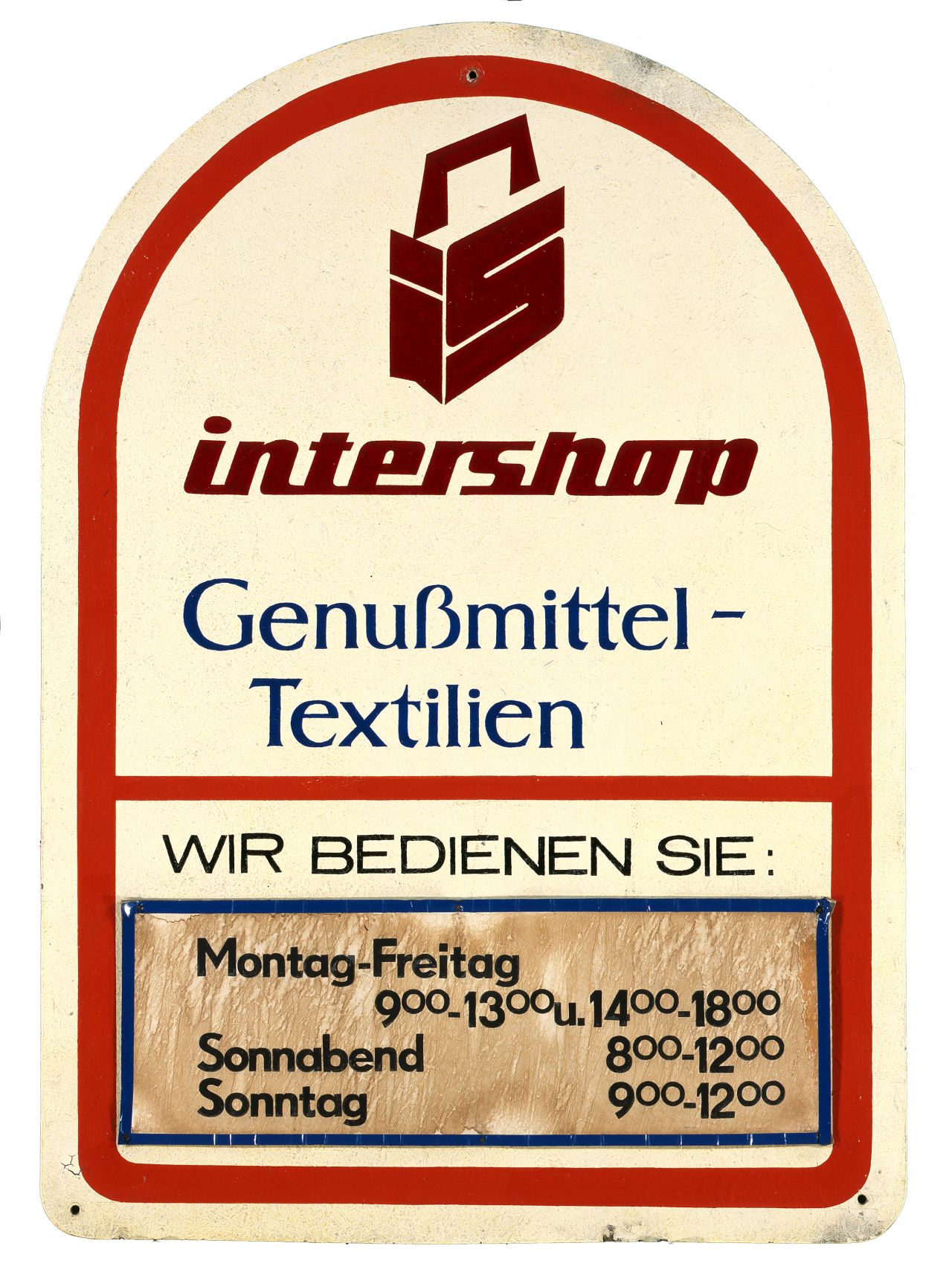 Weißes Holzschild, oben bogenförmig abgerundet, unten gerade. Text: intershop Genußmittel - Textilien  Wir bedienen Sie sowie Öffnungszeiten.