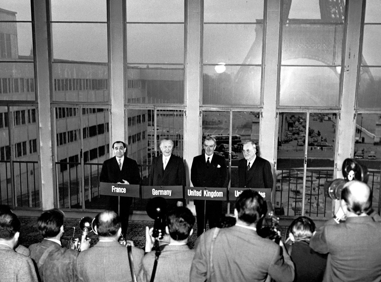 Vier Männer im Anzug (Mendès-France, Adenauer, Eden, Dulles) stehen vor einer Glasfront. Im Hintergrund der Eiffelturm. Vor ihnen jeweils ein Schild mit dem englischen Landesnamen. Ganz vorne: Männer mit Fotoapparaten und einer Kamera von hinten.