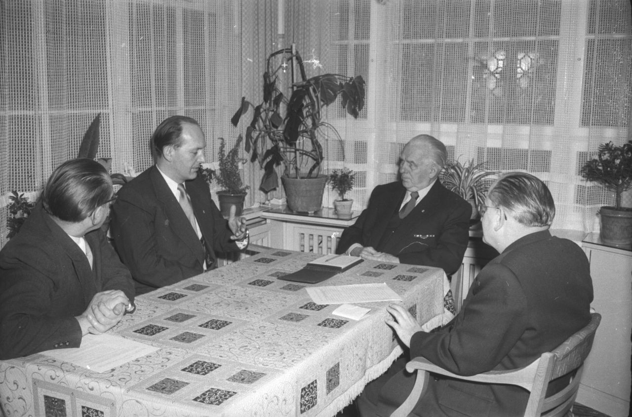 Vier Herren in Anzug, Hemd und Krawatte sitzen an einem Tisch mit Tischdecke. Die beiden mittleren, Pieck und Havemann, sind einander zugewandt; Havemann spricht. Im Hintergrund eine Fensterbank mit Topfpflanzen und Netzgardinen.
