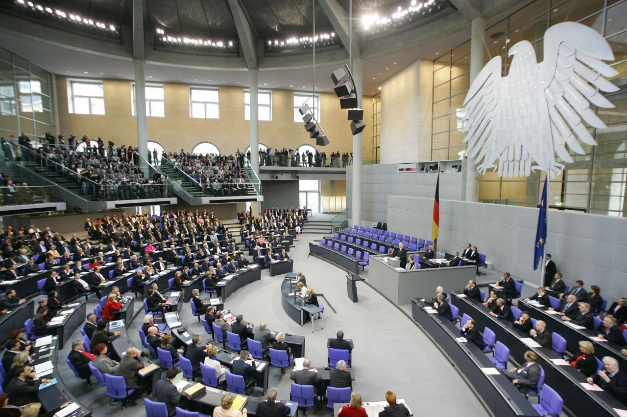 Seitlicher Blick von oben, Plenarsaal des Bundestages. Rechts an der Wand ein großer Bundesadler, darunter die deutsche und europäische Flagge und vier Sitzreihen. Gegenüber im Halbrund Reihen mit den restlichen Abgeordneten. Oben links Besuchertribünen.