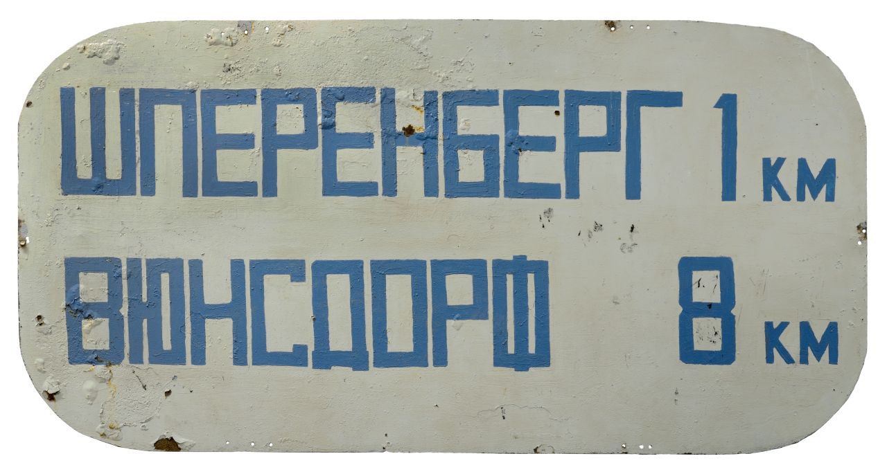 Silbergraues Schild mit gerundeten Ecken. Auf der Vorderseite in blauer, kyrillischer Schrift zwei Namen mit Kilometerangaben (1 km, 8 km).