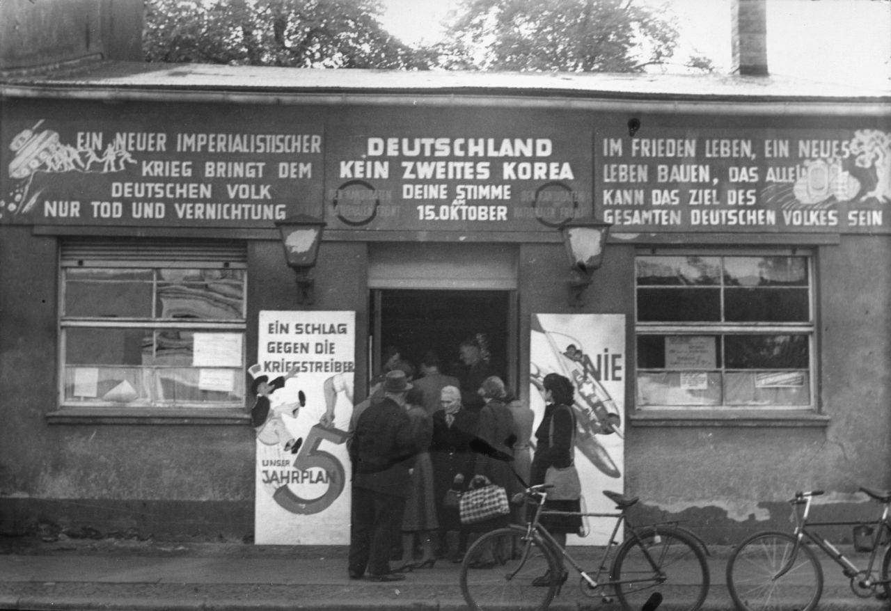 Schwarzweißfoto; flaches Gebäude, vor und im Eingang wartende Menschen. An der Fassade hängen Schilder mit Wahlkampfparolen. Zwei Herrenfahrräder sind vor dem Bürgersteig abgestellt.