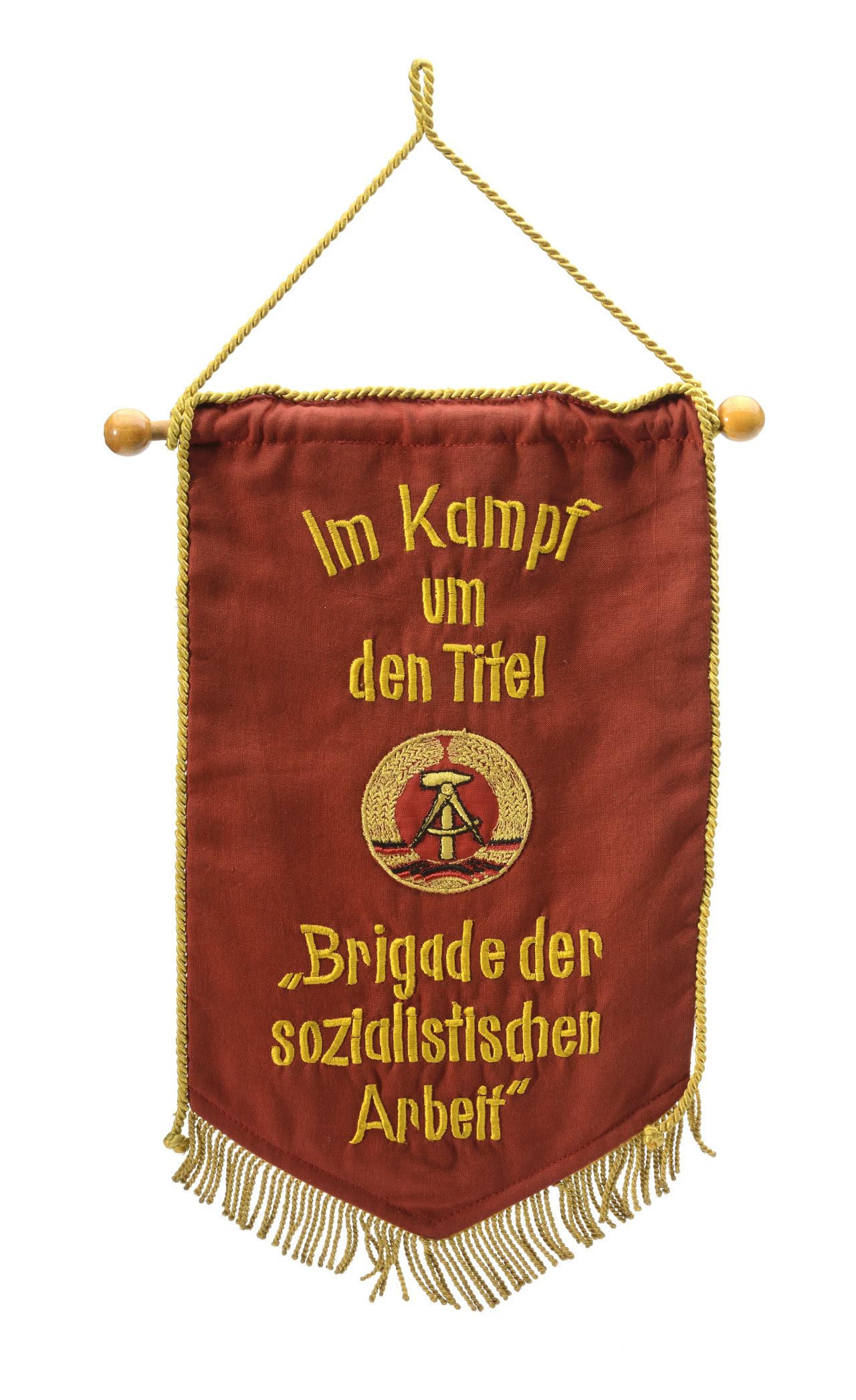 Roter Wimpel umlaufend mit goldfarbener Fransenkordel, beide Seiten unterschiedlich bestickt. Auf dieser Seite Text: Im Kampf um den Titel, darunter DDR-Wappen, darunter Text: 'Brigade der sozialistischen Arbeit'