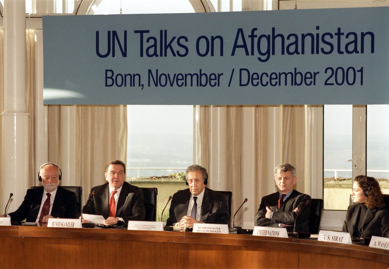 Farbfoto; in einer Reihe sitzend: Francesc Vendrell, Gerhard Schröder, Lakhdar Brahimi, Joschka Fischer und Ms. Zia. Über ihnen ein Schild mit Schriftzug 'UN Talks on Afghanistan, Bonn, November/December 2001'. Dahinter mit Vorhängen umrahmte Fenster.