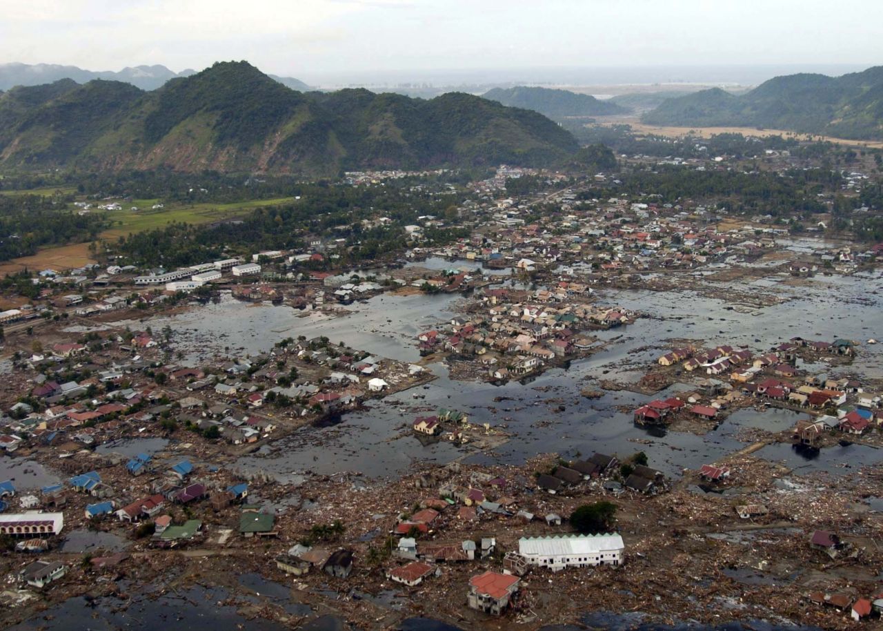 Farbige Luftaufnahme von einem durch den Tsunami überschwemmten und fast völlig zerstörten Dorf. Vereinzelt stehen noch Häusergruppen, drum herum befinden sich nun Schutt- und überschwemmte Flächen. Im Hintergrund bergiges, grünbewachsenes Umland.
