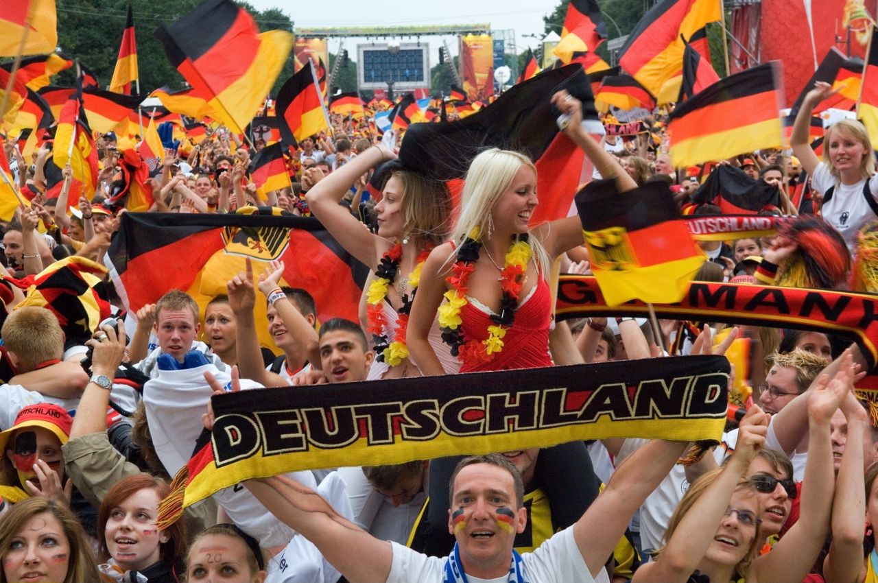 Farbfoto; jubelnde Fußball-Fans auf der Fanmeile; einige haben die deutschen Farben aufs Gesicht gemalt, viele mit Deutschlandfahnen und Accessoires in deutschen Farben. Fan im Vordergrund hält schwarz-rot-gelben Schal mit Lettern 'Deutschland' hoch.