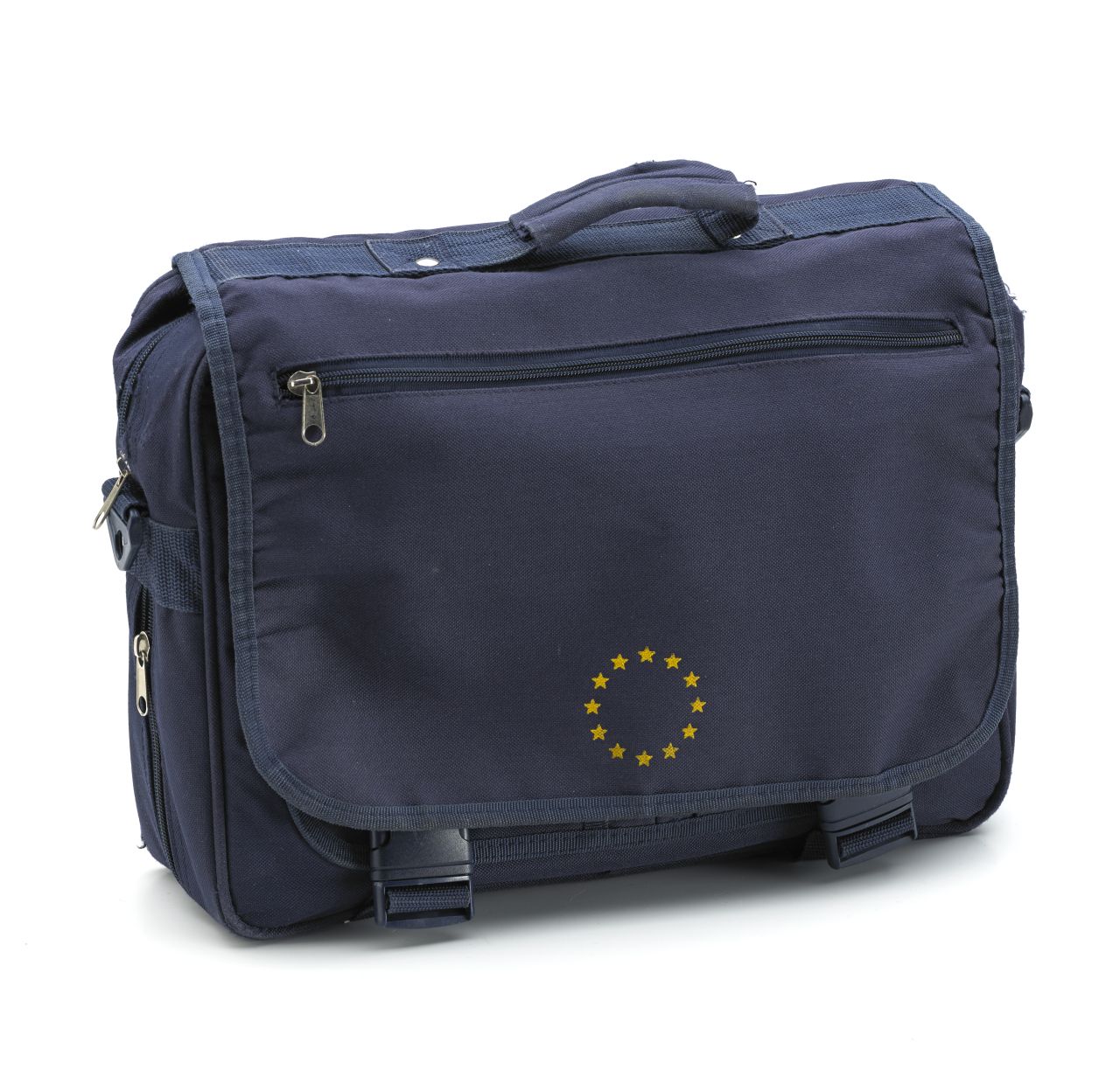 Blaue Tasche mit Reißverschluss. Darüber blaue Taschenklappe mit zwei Steckverschlüssen und Sternenkranz aus 12 gelben Sternen unten. Auf der Innenseite der Klappe gelb: European Conference on Company Taxation / Brussels, 29 and 30 April 2002. Auf der Tasche unter der Klappe verschiedene Einsteckfächer. Seitlich und unten ein weiterer Reißverschluss. Mit Tragegriff und Trageriemen.