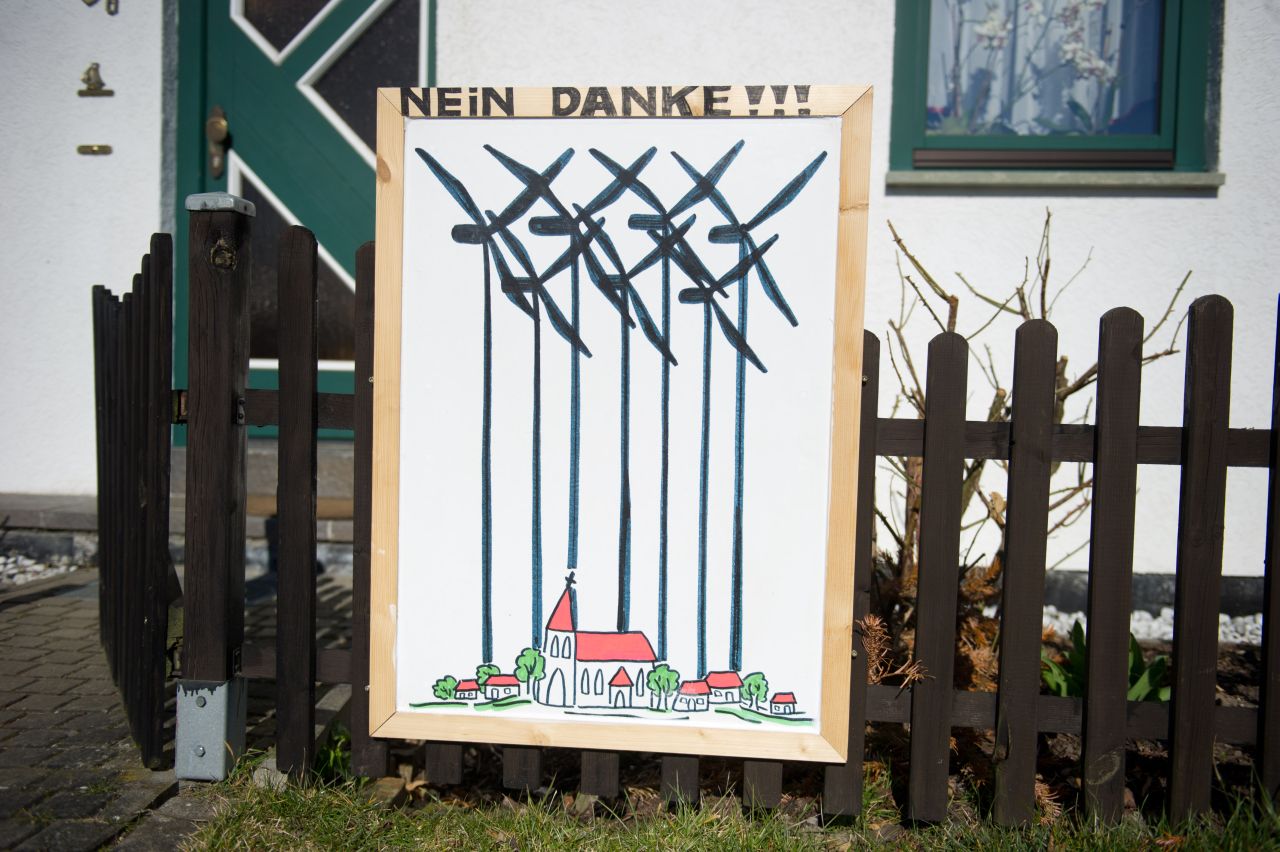 Auf dem Foto ist ein Plakat in einem Vorgarten zu sehen. Das Plakat hängt an einem Holz-Gartenzaun. Auf diesem ist eine Zeichnung zu sehen: Ein idyllisches Dorf mit Kirchturm als höchstem Gebäude. Über diese gezeichnete Idylle sind in schwarzen, dicken Pinselstrichen moderne Windräder gemalt. Die parallelen Türme der Windräder über dieser Idylle wirken wie Gitterstäbe eines Käfigs, die das Dorf umgeben.