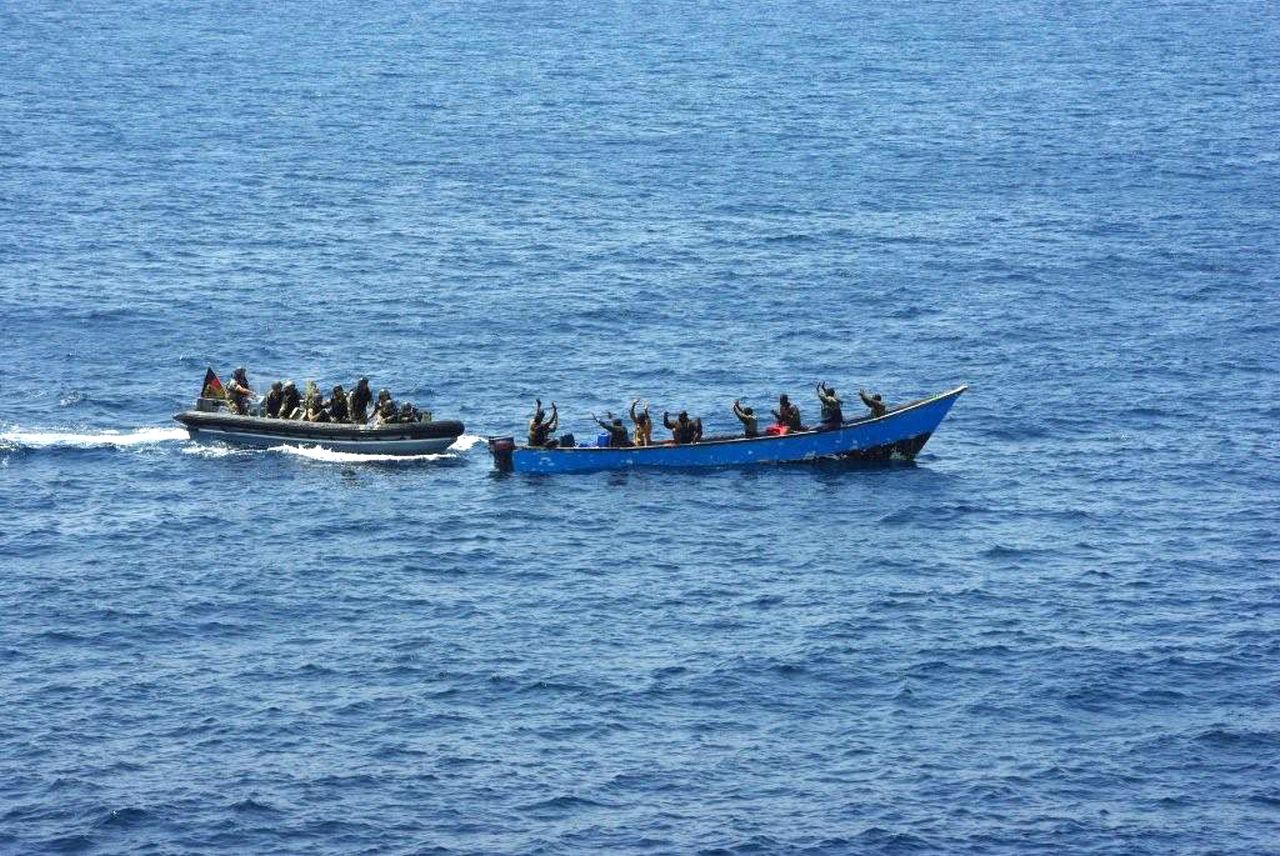 Das Foto zeigt zwei Boote mitten im Meer. Das Wasser ist tiefblau und sehr ruhig. Ein Boot voller Soldaten fährt an ein Piraten-Boot heran. Acht Piraten sind zu erkennen, die alle die Hände hochheben. Am Bundeswehr-Boot ist eine deutsche Fahne zu erkennen. Die Soldaten tragen alle Uniformen und Ausrüstung.