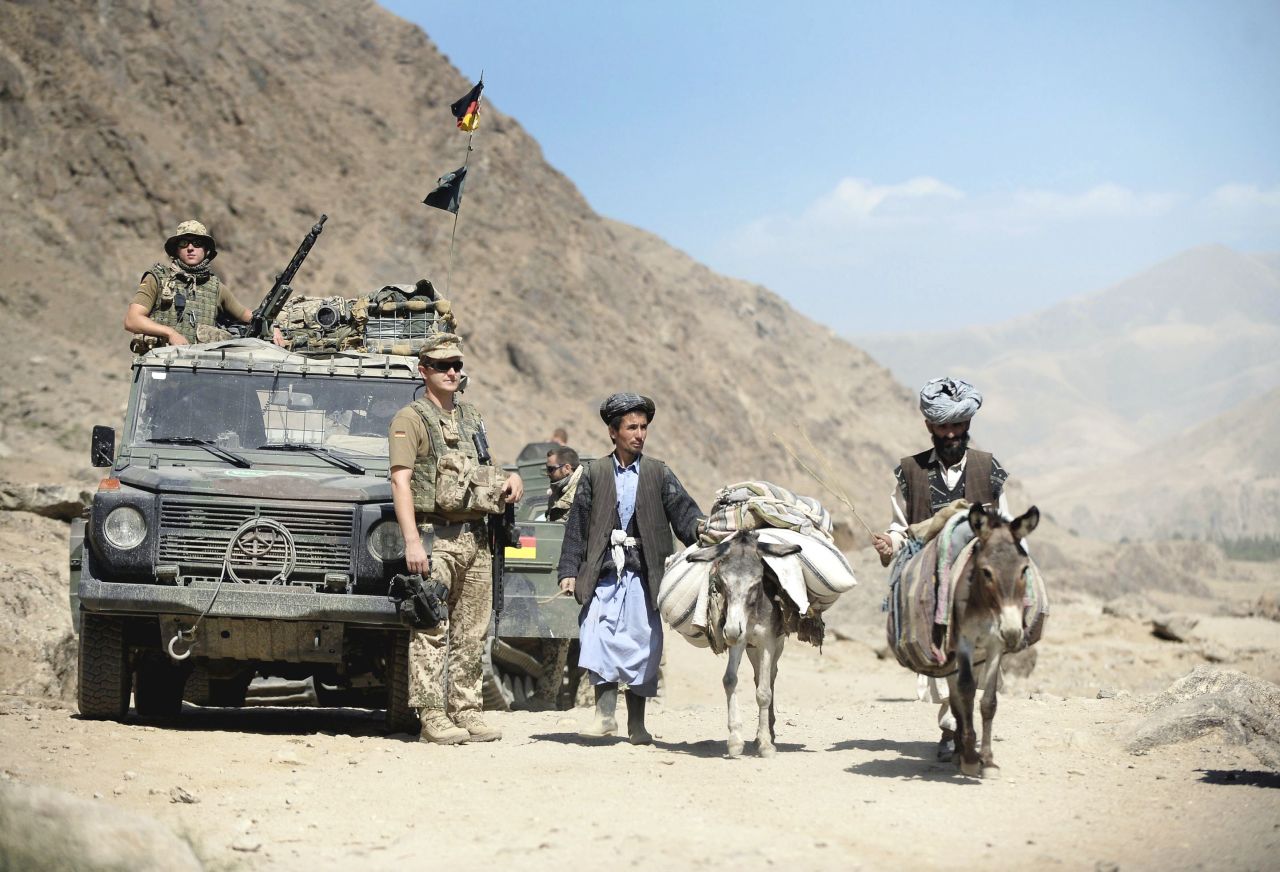 Das Foto zeigt links drei Bundeswehrsoldaten auf Patrouille in Afghanistan. Sie stehen vor, auf und neben einem Militär-Geländewagen. Rechts im Bild sind zwei afghanische Zivilisten zu sehen, die mit zwei bepackten Lasteseln unterwegs sind. Im Hintergrund sieht man unbewachsene Berge und blauen Himmel. 