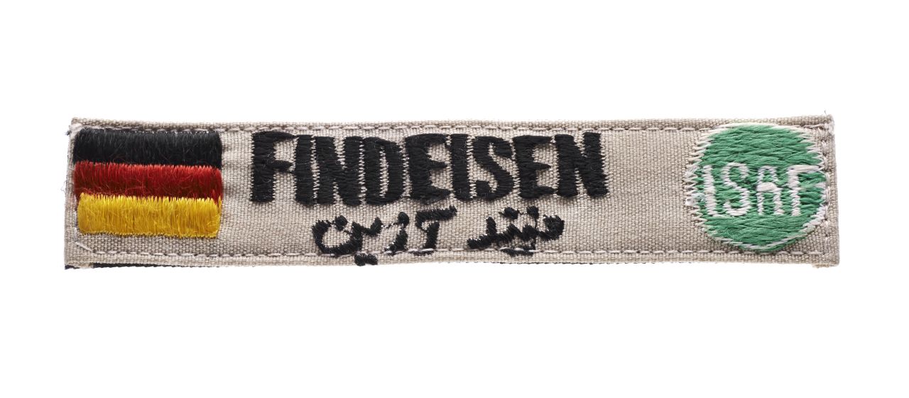 Auf einem khakifarbenen, rechteckigen Stück Stoff ist der Name des deutschen ISAF-Soldaten Harald Findeisen mit schwarzem Faden aufgenäht, sowohl in lateinischen als auch in arabischen Buchstaben. Links ist eine Deutschlandflagge aufgenäht. Rechts sieht man das runde ISAF-Logo.