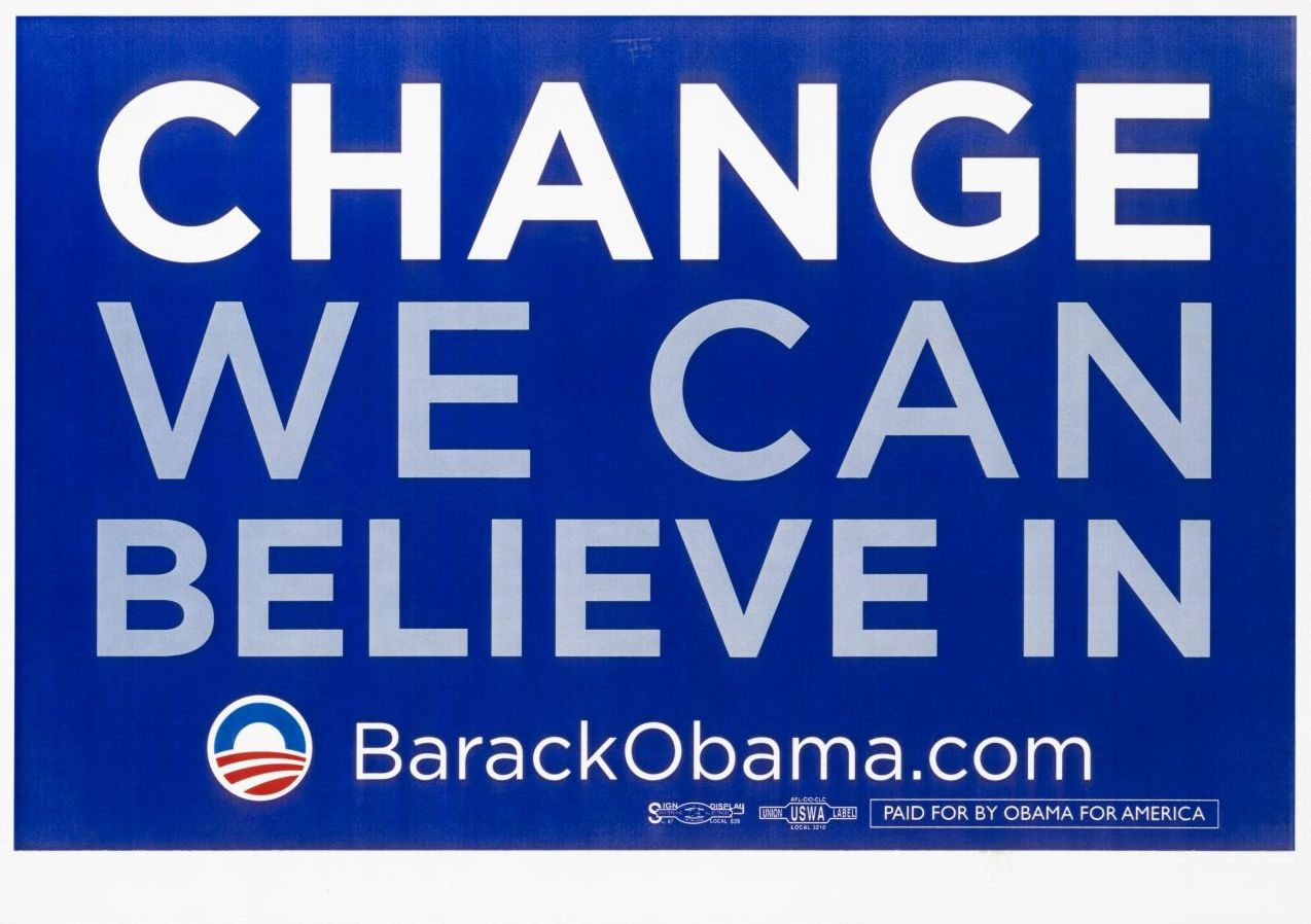 Querformatiges laminiertes Plakat; dunkelblauer Grund mit weißem Rahmen; mittig in weißen und grauen Großbuchstaben 'Change we can believe in'; unten links Logo der Kampagne 'Obama for America' in blau und rot; daneben in weißer Schrift 'Barack Obama.com'. 