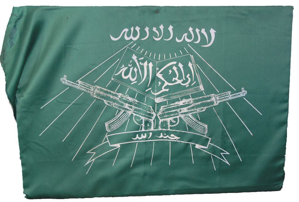 Grünes Tuch mit arabischen Schriftzeichen über einem Koran und gekreuzten Gewehren vom Typ Kalaschnikow. Unten Schriftband mit arabischem Text. Im Hintergrund stilisierte Sonne. 
Ursprünglich auf einer braunen Platte befestigt.