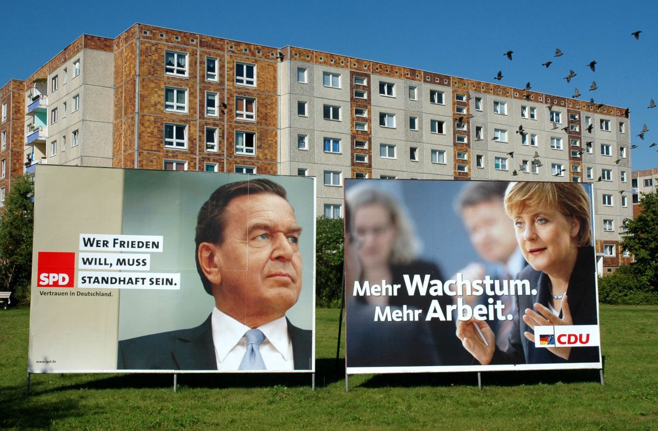 Farbfoto von zwei Plakatwänden vor Hochhäusern. Links Plakat SPD mit Foto Gerhard Schröder und Logo SPD, schwarzer Aufschrift: Wer Frieden will, muss standhaft sein. Daneben Plakat CDU mit Foto Angela Merkel und Logo CDU, weiße Aufschrift: Mehr Wachstum. Mehr Arbeit.