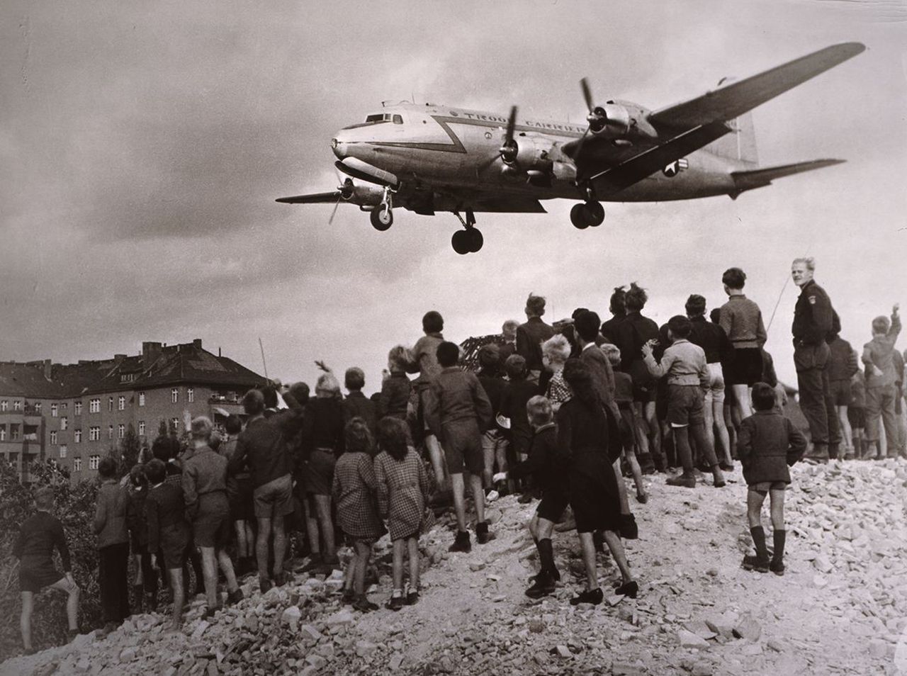 Eine Gruppe von Menschen, auf einem Schuttberg stehend, schauen einem im Landeanflug befindlichen Rosinenbomber entgegen. Links im Hintergrund ein mehrstöckiges Haus