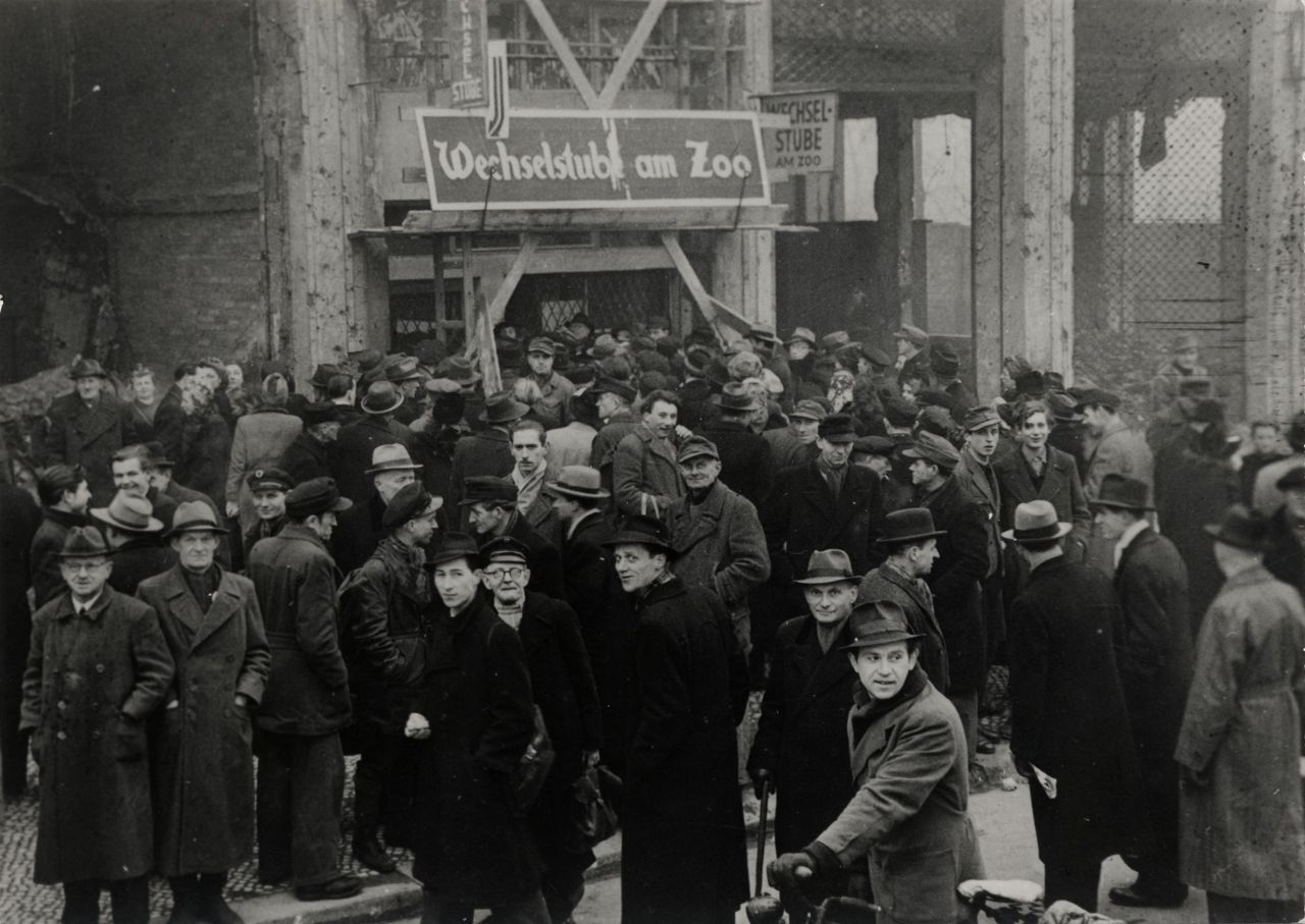 Schwarz-Weiß-Foto einer größeren Menschenmenge, vorwiegend Männer, wenige Frauen vor einer Wechselstube. Fast alle tragen Mantel und Hut. Zwei Schilder oberhalb des Eingangs zeigen den Namen  'Wechselstube am Zoo' an.