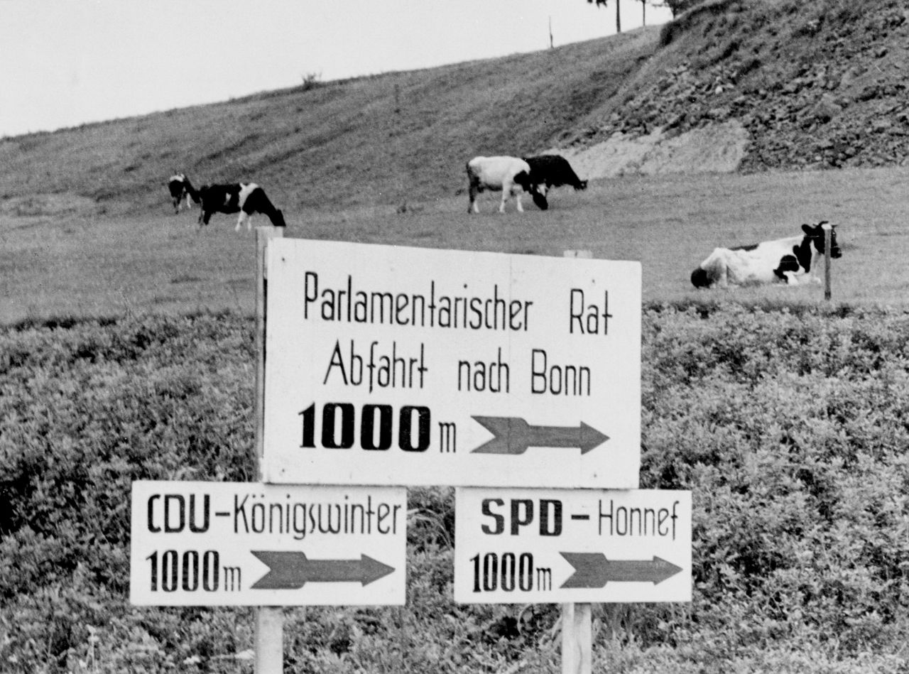Schwarz-Weiß-Foto: Im Vordergrund 3 Schilder an 2 Pfählen befestigt. Schriftzüge darauf: oben 'Parlamentarischer Rat Abfahrt nach Bonn 1000m', links unten 'CDU-Königswinter 1000m', rechts unten 'SPD-Honnef 1000m', im Hintergrund Hang mit einer Kuhweide.