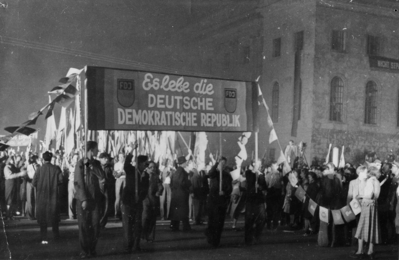 Schwarz-weiße Abbildung eines Demonstrationszuges während des Abends. Im Vordergrund ragt ein Transparent mit der Beschriftung: FDJ Es lebe die Deutsche Demokratische Republik FDJ , auf. Dahinter laufen sehr viele Fahnenträger.