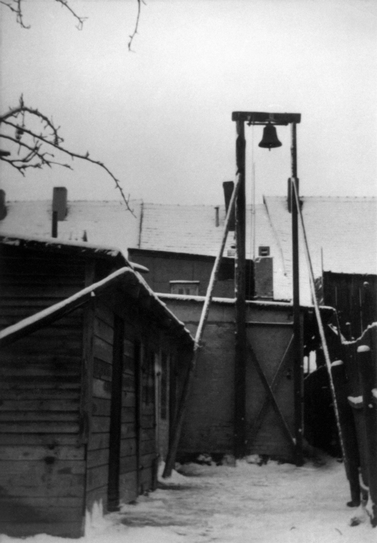 Schwarz-weißes Foto-Positiv mit der Abbildung einer Holzbaracke im Vordergrund, neben der an einem hohen Holzgerüst eine Glocke hängt. Im Hintergrund weitere Gebäude.