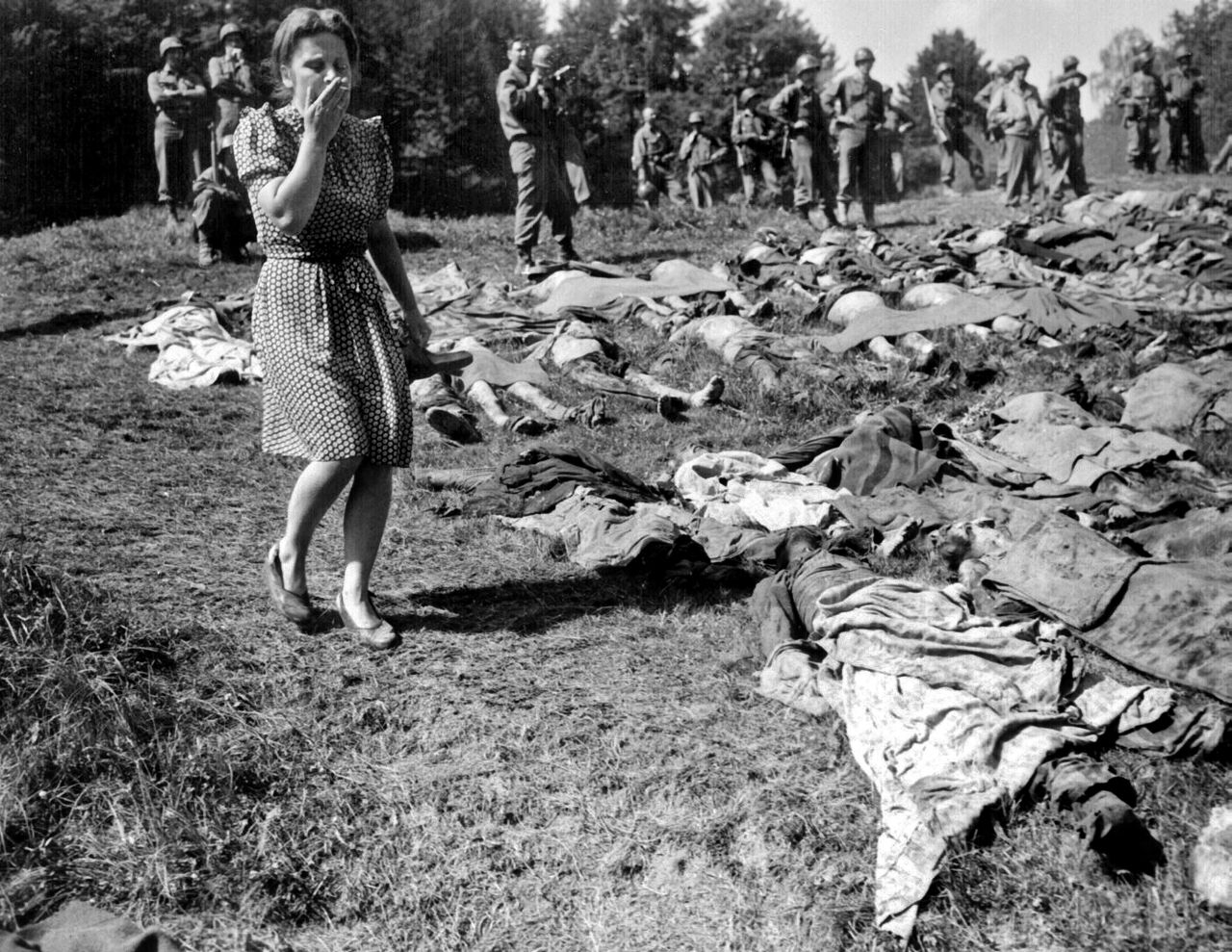 Im bayerischen Nammering betrachtet die Bevölkerung mit Scham und Entsetzen hunderte Leichen von Häftlingen des KZ Dachau. Das Foto zeigt viele Leichen, die nebeneinander auf dem Boden liegen und mit Stoffstücken teilweise bedeckt worden sind. Neben den Leichen steht eine Frau, die mit einer Hand ihre Nase und ihren Mund bedeckt. Im Hintergrund stehen Soldaten und betrachten die Frau und die Leichen.