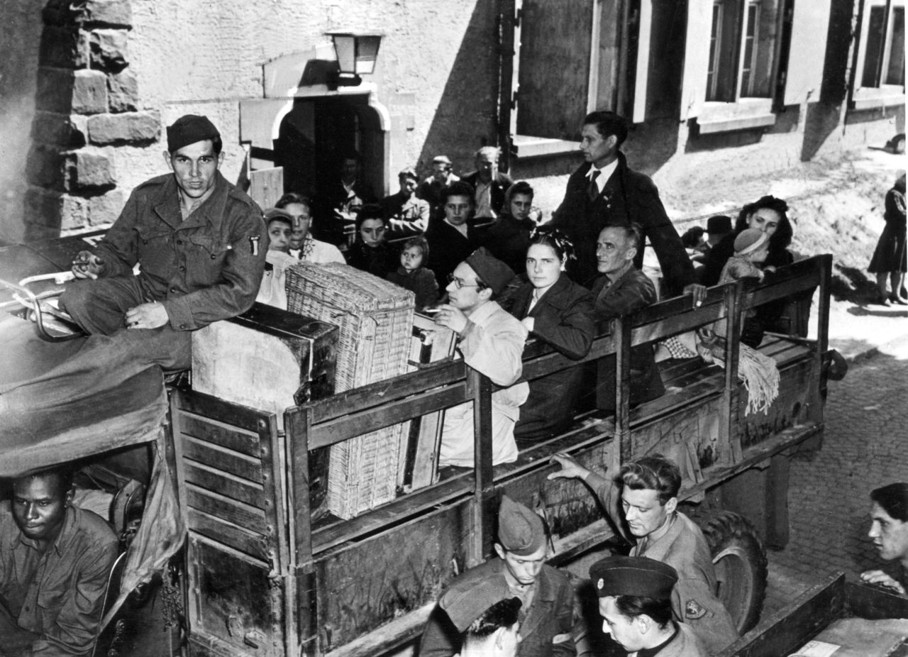 Eine Gruppe von Menschen sitzt auf der offenen Ladefläche eines kleinen Lastautos. Links ein Stück des Fahrerhäuschens mit Fahrer. Oben erhöht gegenüber den anderen Personen ein Mann in Uniform. Seitlich des Lastautos weitere Menschen.