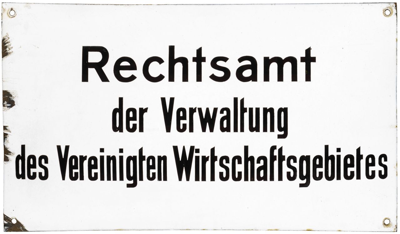 Weißes, gewölbtes Schild; Text (schwarz): Rechtsamt der Verwaltung des Vereinigten Wirtschaftsgebietes. In den Ecken jeweils ein Loch zur Befestigung.